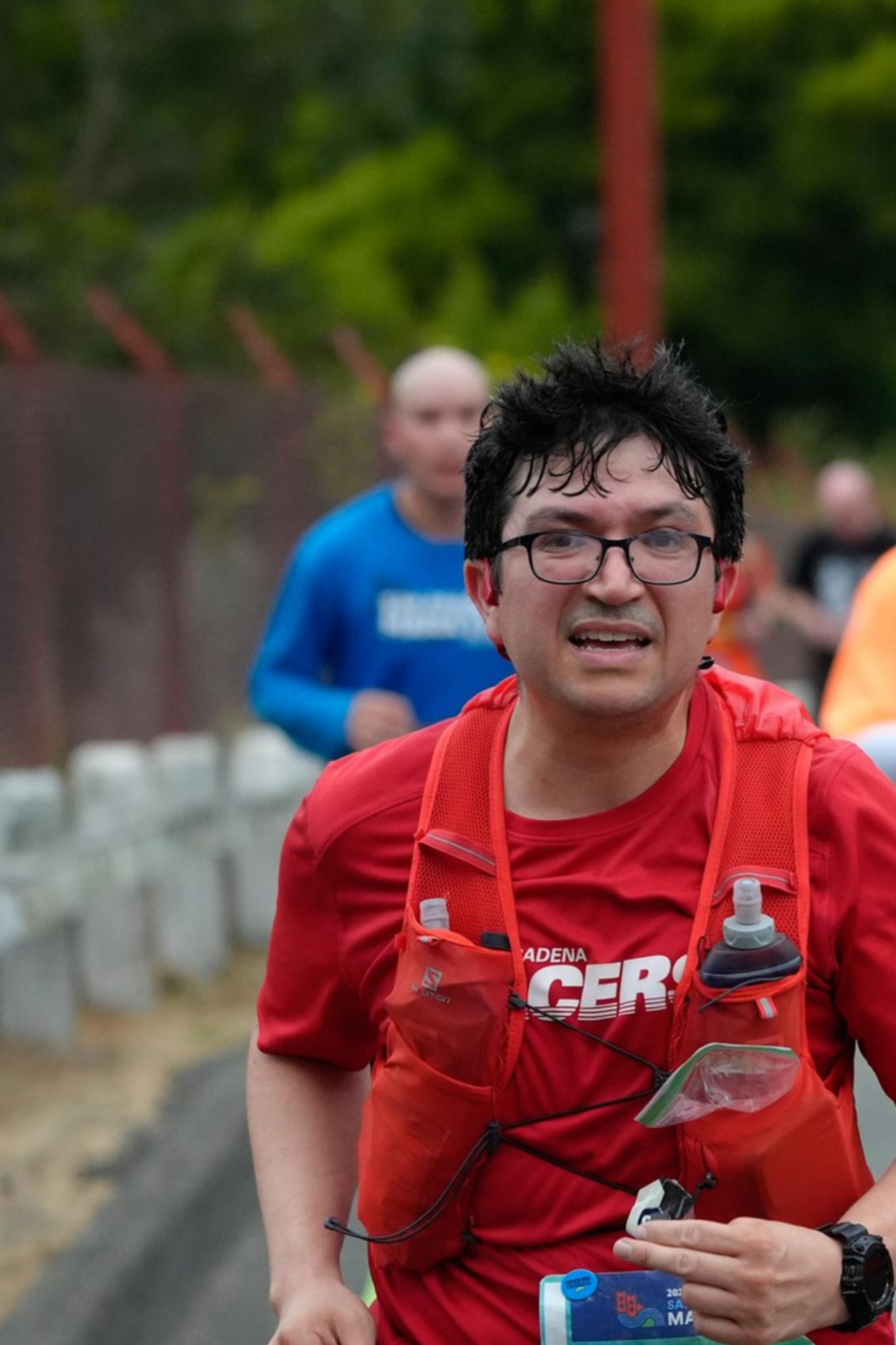 A man wearing a red shirt, glasses, and a water bottle vest runs in a marathon with other runners in the background on a road lined with traffic cones. 