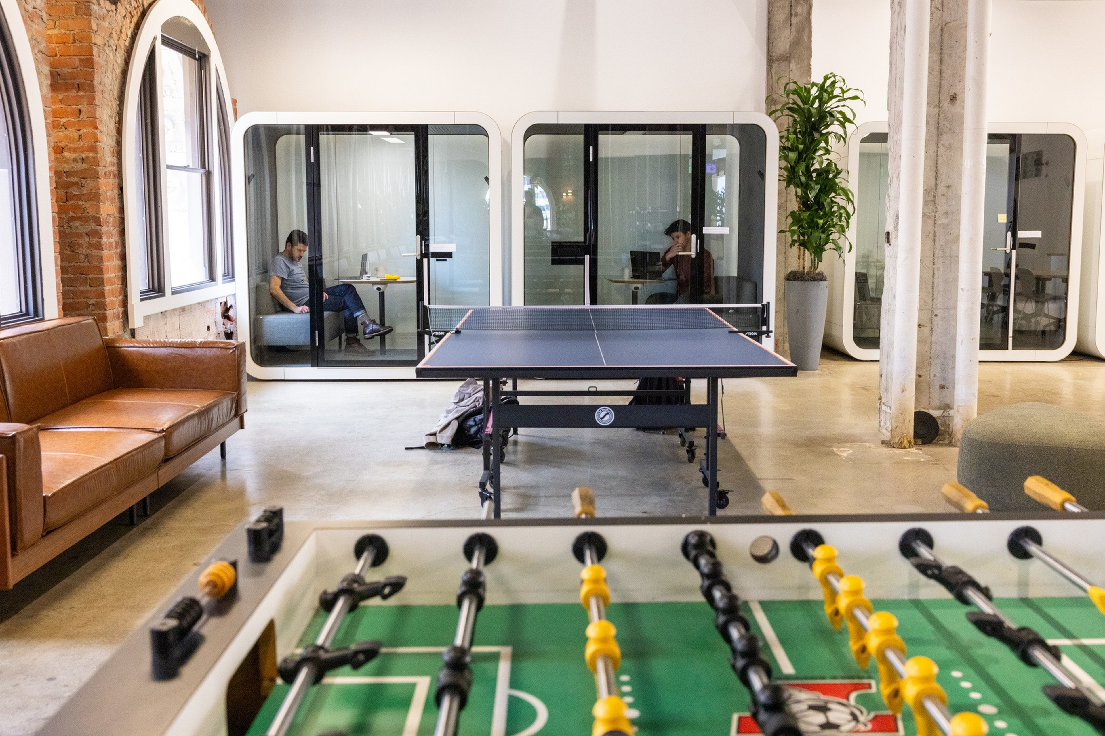 A modern office lounge contains a foosball table, a ping pong table, a brown leather sofa, and two people sitting alone in glass pods.