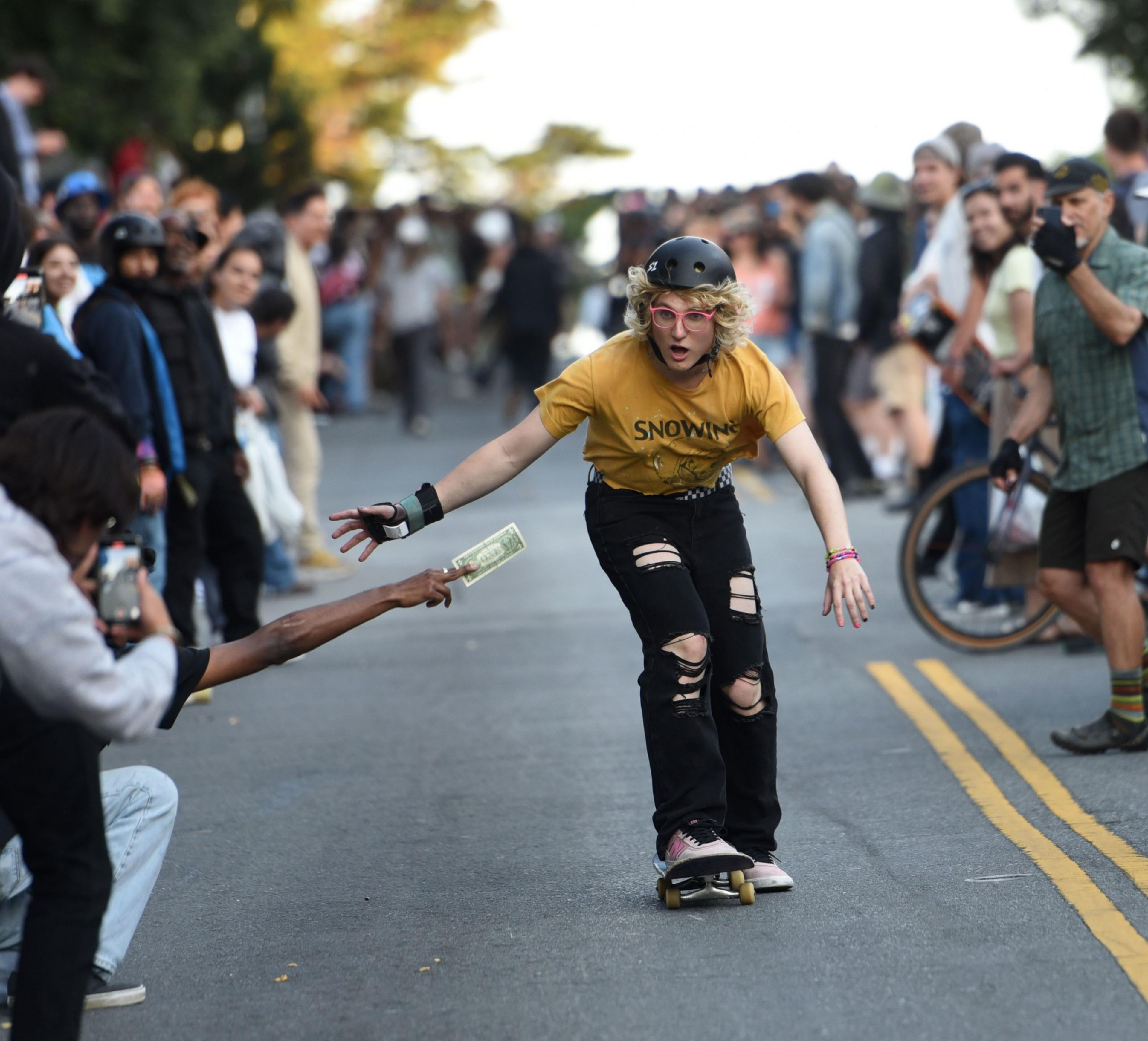 A skater in a yellow shirt and helmet rides down a street lined with spectators, reaching for a dollar bill held out by a kneeling person taking a photo.
