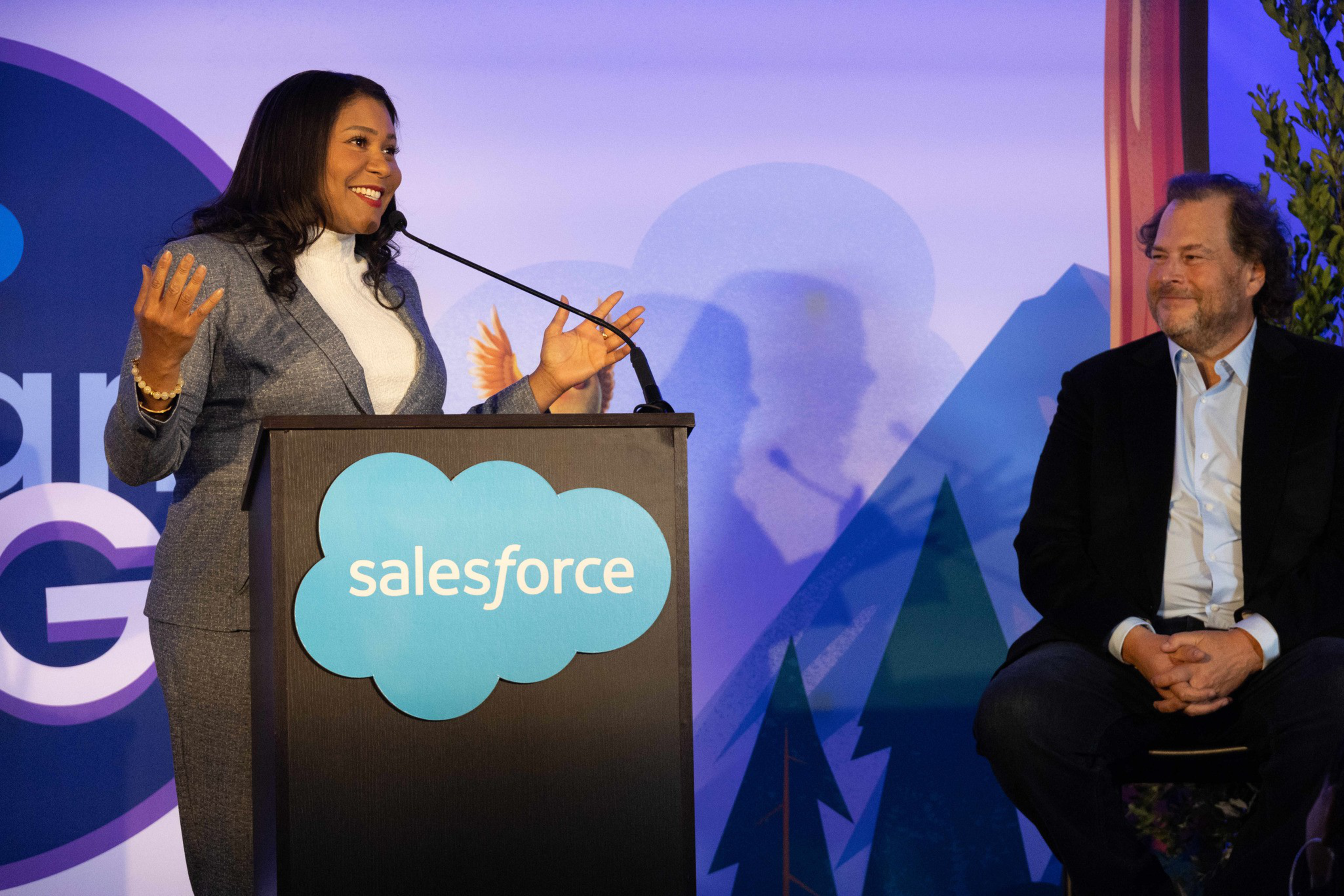 A woman in a grey suit speaks at a podium with a Salesforce logo, while a man in a black jacket and light shirt sits smiling nearby, in front of a blue and purple backdrop.