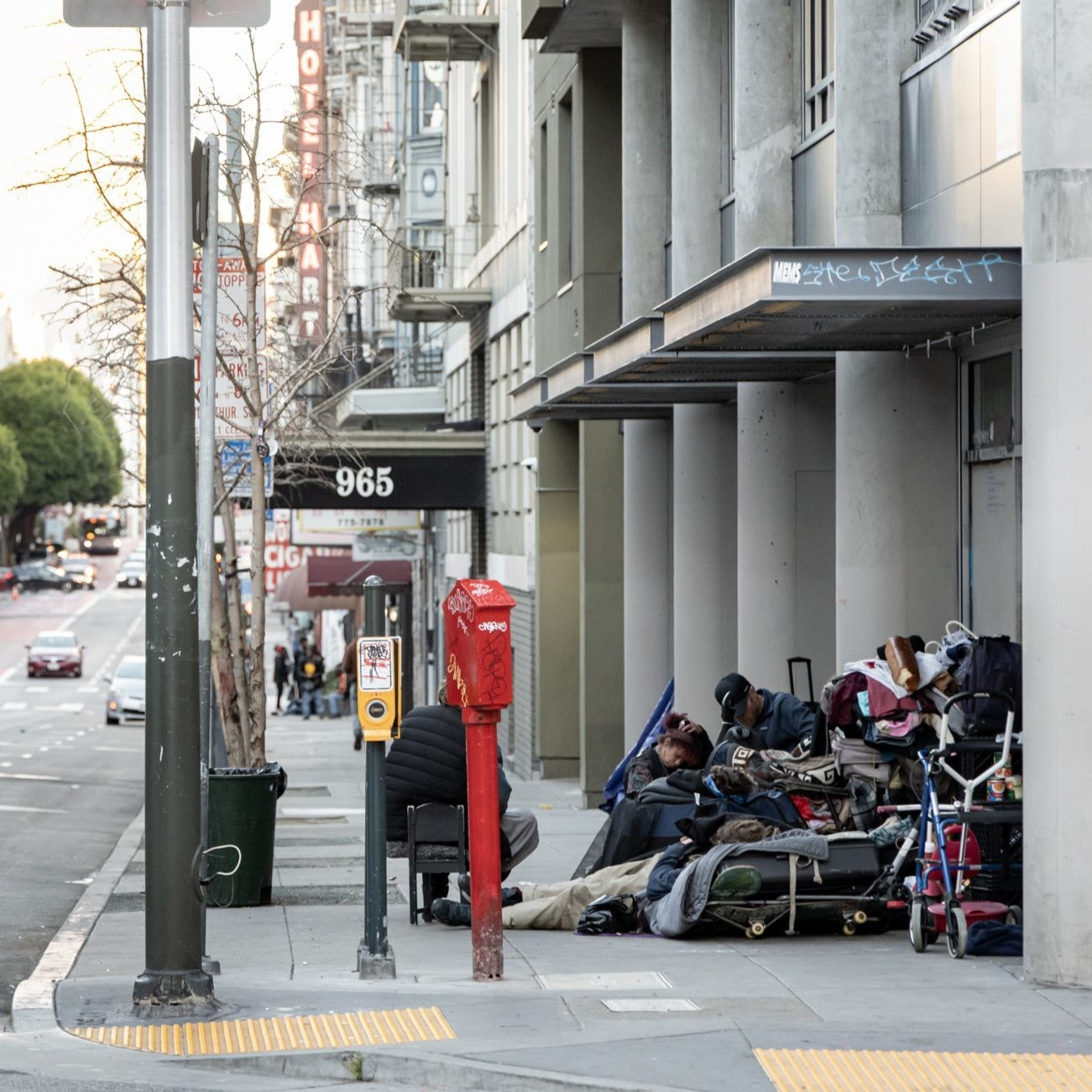 A city street scene shows people sitting amidst belongings like bags and carts. There's a hotel sign in the background under a cloudy sky.