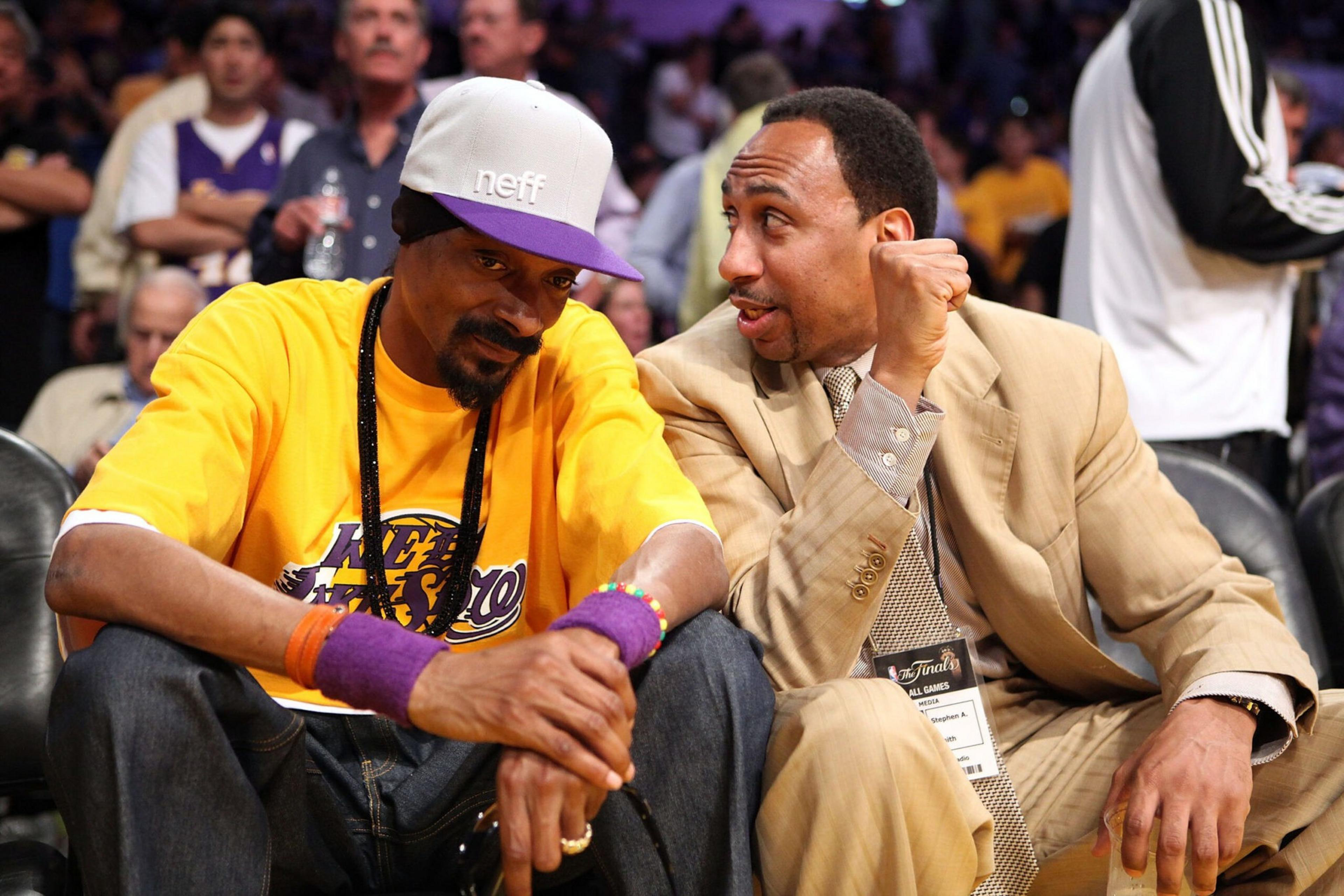 A man in a yellow Lakers shirt and white cap sits next to another man in a beige suit who is talking animatedly. They appear courtside at a basketball game.
