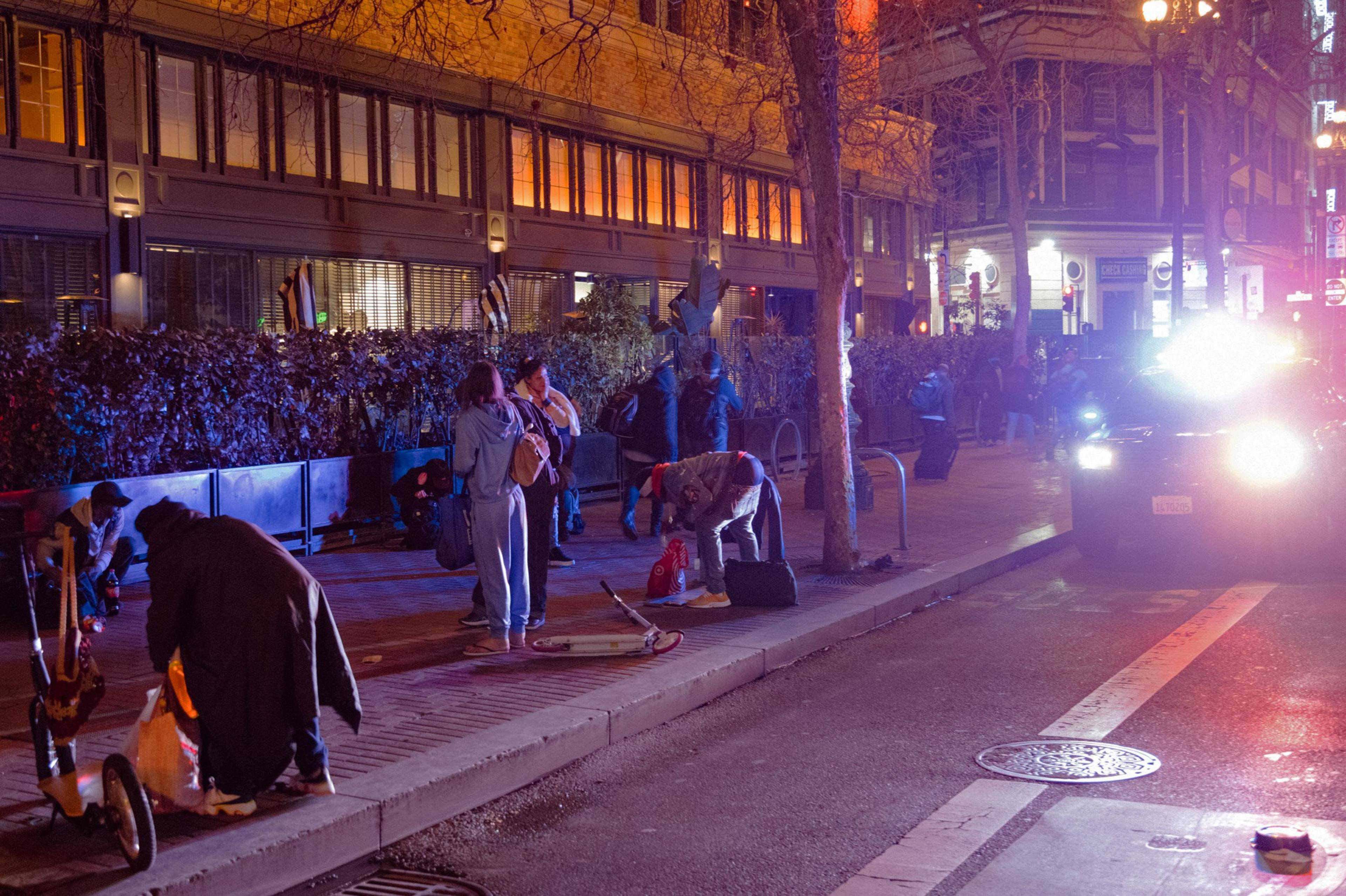 Night scene on a city street with individuals and bright lights from an oncoming car.