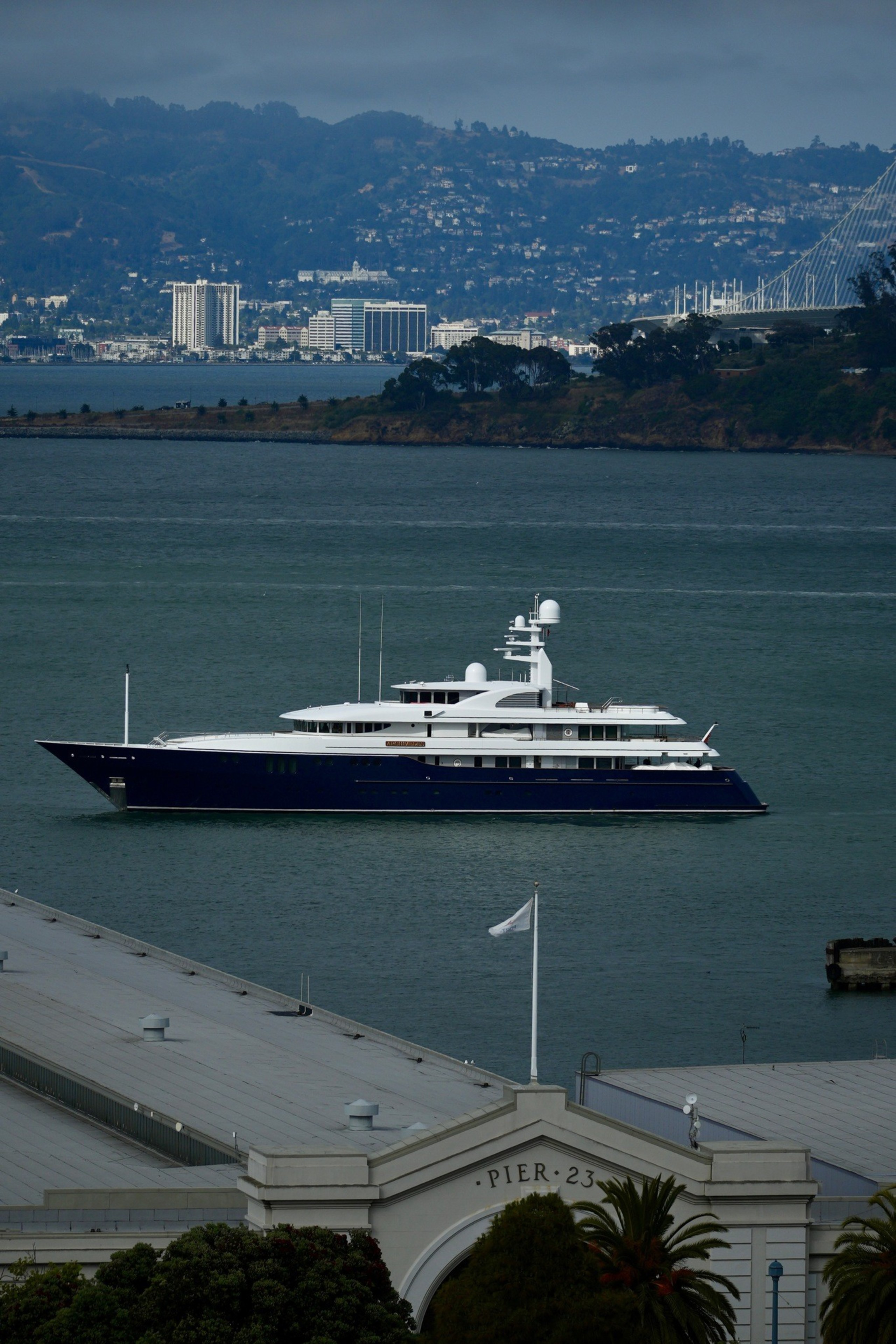 A large, sleek yacht with a dark hull and white superstructure sails near a waterfront labeled "Pier 23," with hills and buildings in the background.