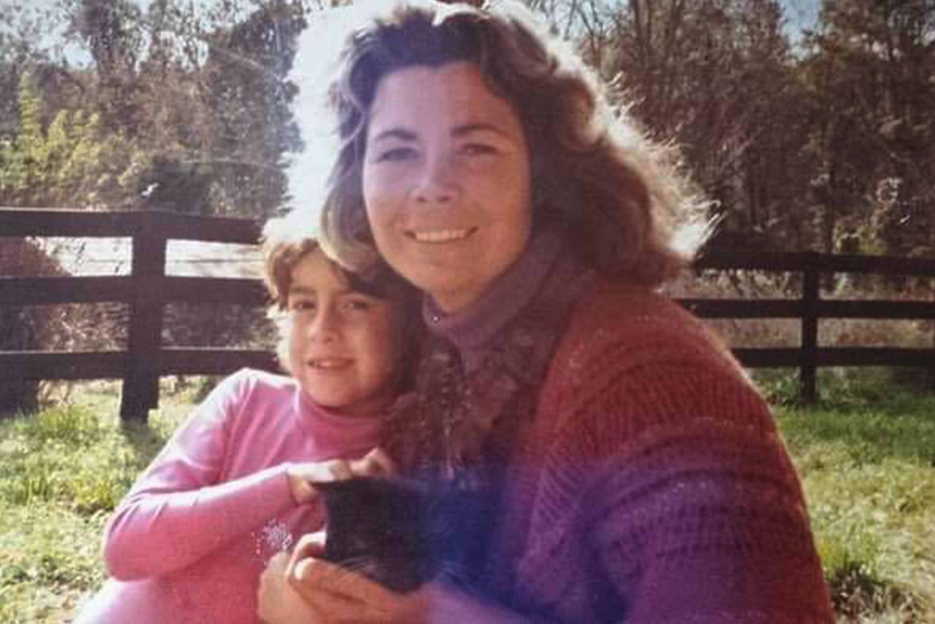 A woman and a girl sit closely together outside near a wooden fence, smiling warmly while holding a small black kitten.