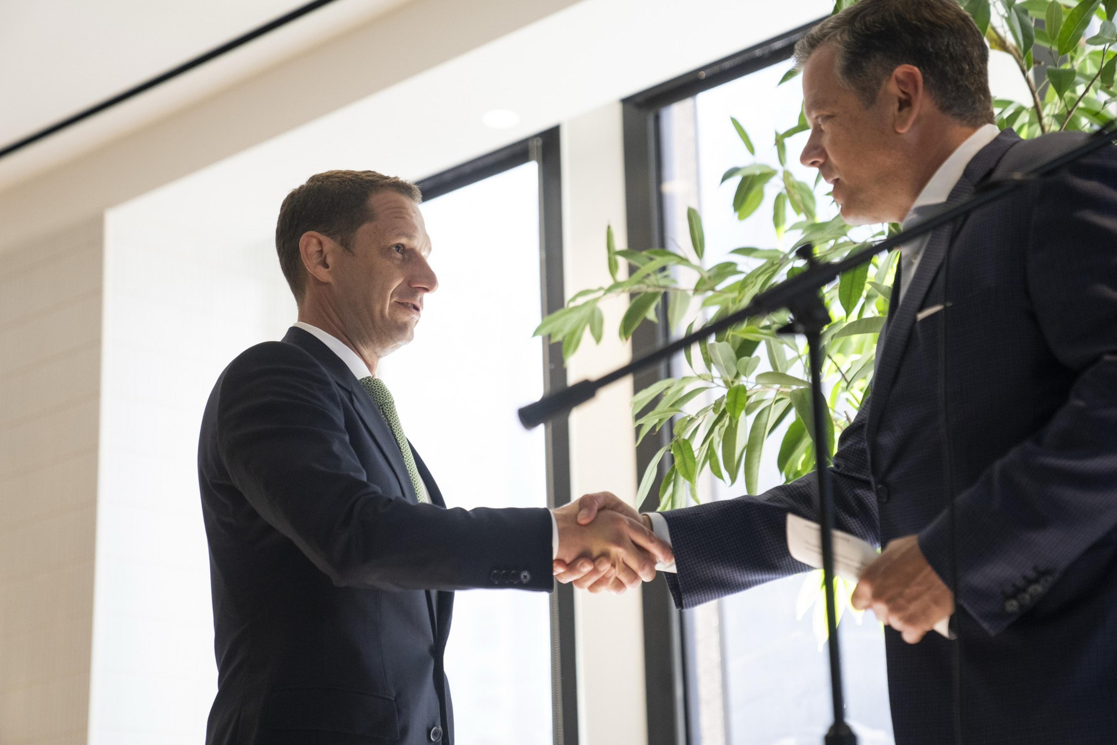 Two men in suits shake hands indoors near a window, with a leafy plant in the background. A microphone is in the foreground.