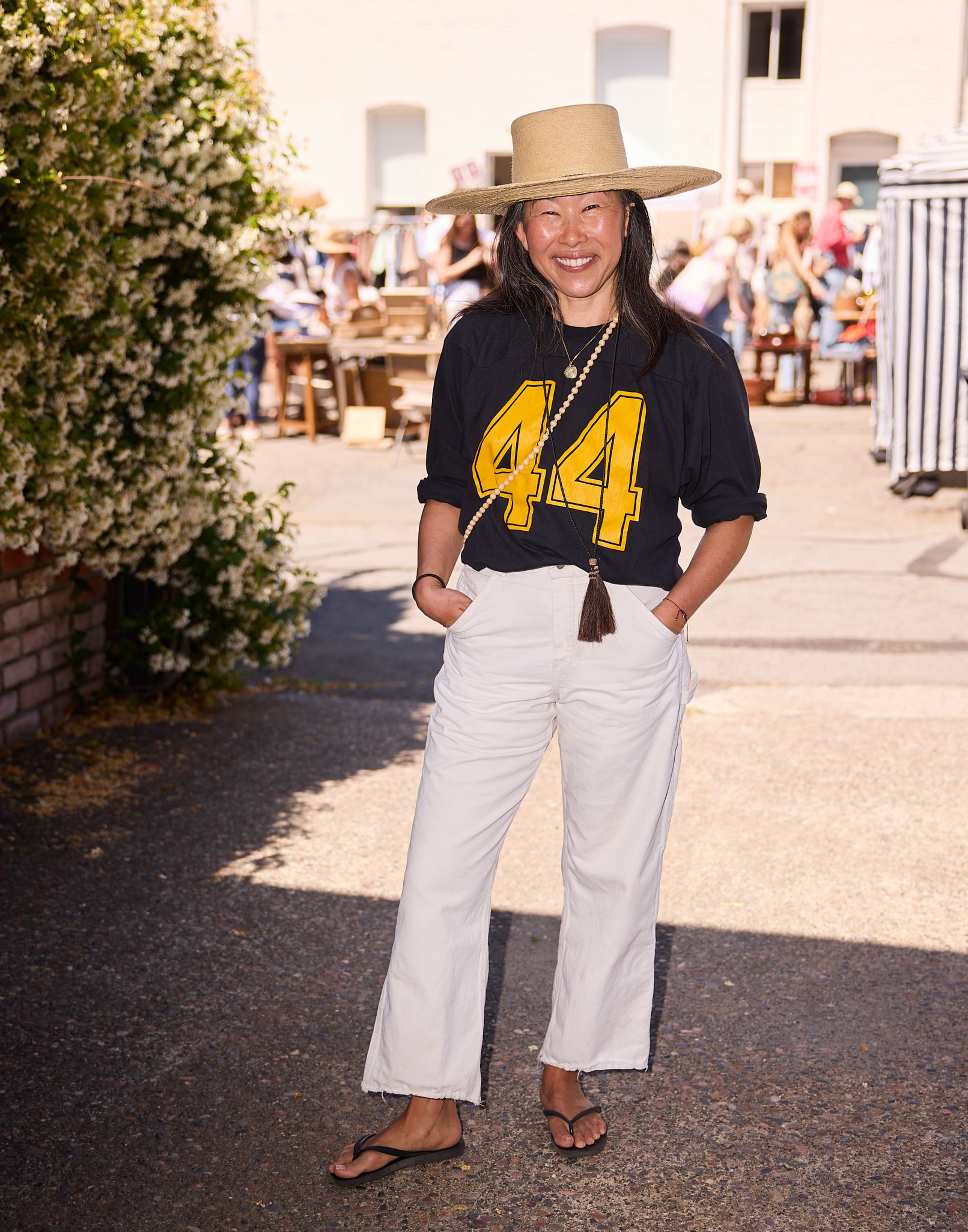A woman smiles wearing a large straw hat, black shirt with yellow “44,” white pants, and sandals, standing outdoors near bushes and people.