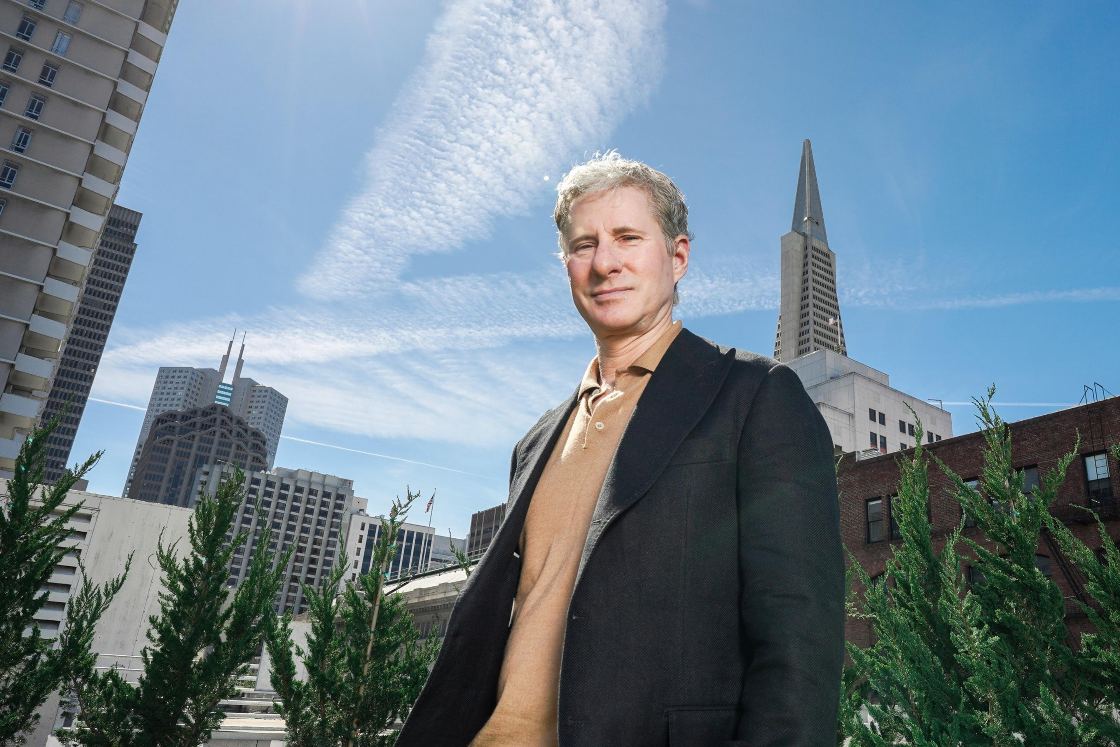 A man in a black jacket stands outside, with skyscrapers and clear blue sky featuring wispy clouds in the background. Green foliage is in the foreground.