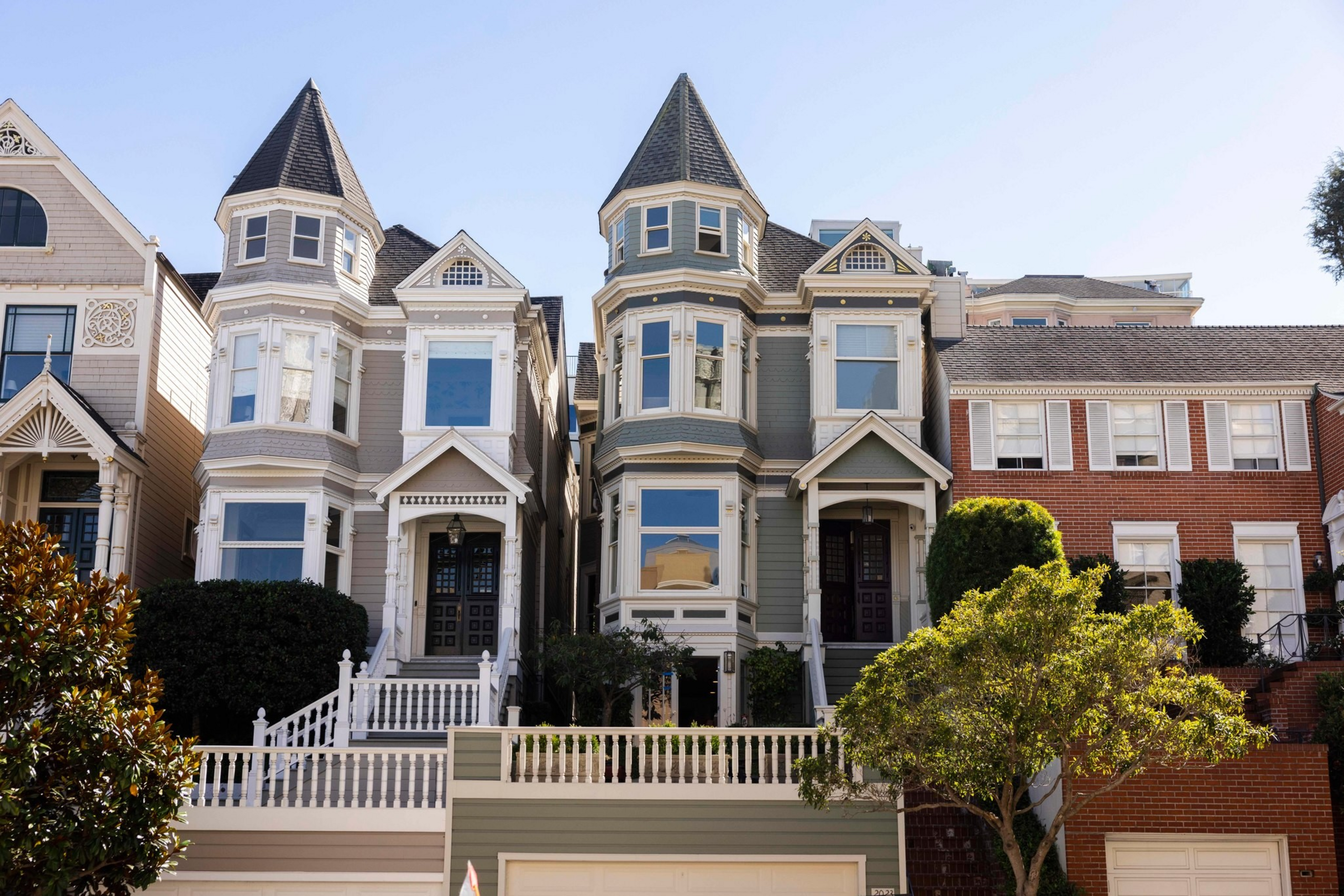 Two Victorian-style homes with pointed turrets, large windows, and front porches stand side by side, flanked by brick buildings and greenery.