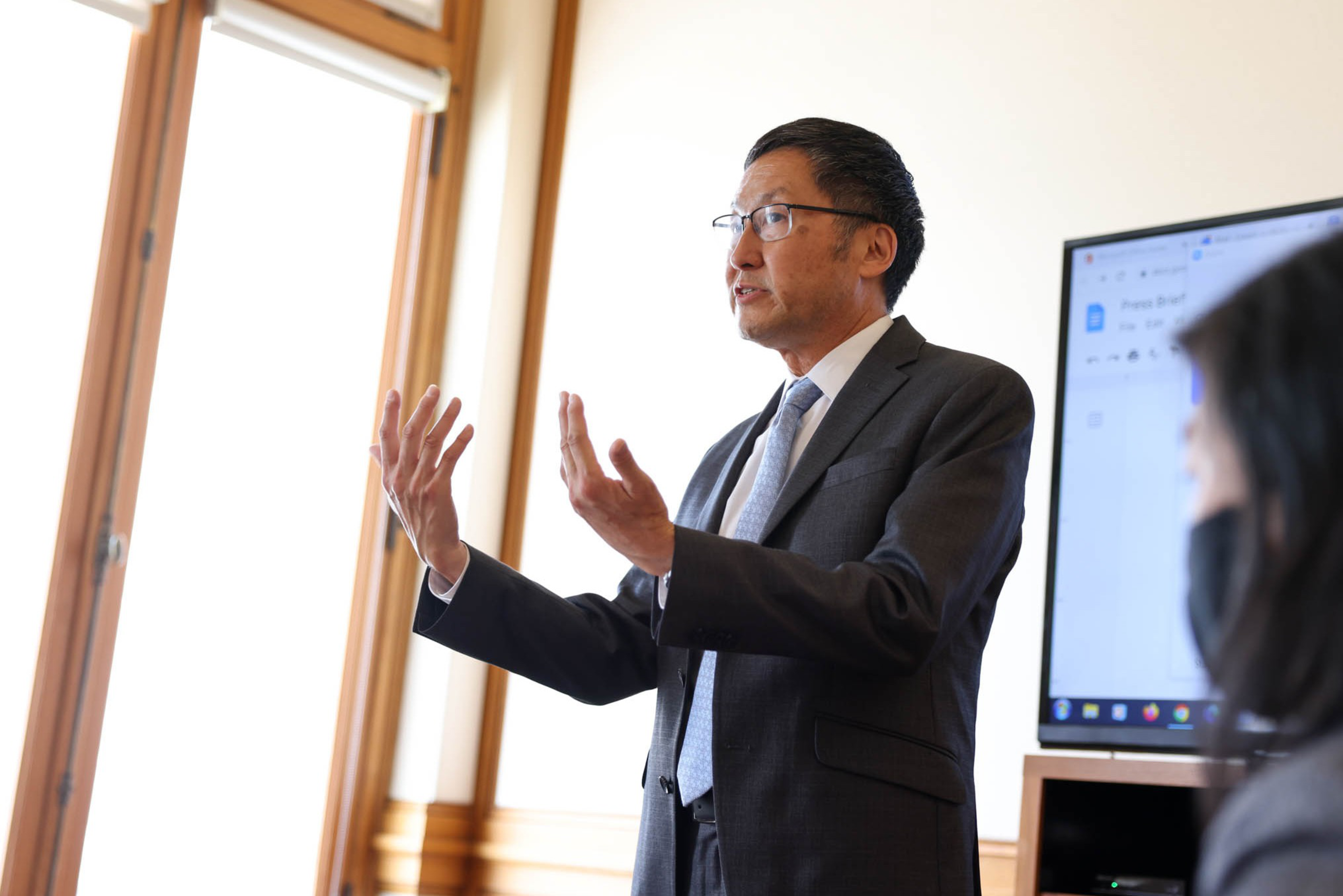 A man in a suit and tie is speaking and gesturing with both hands in a room with large windows and a screen displaying a document.
