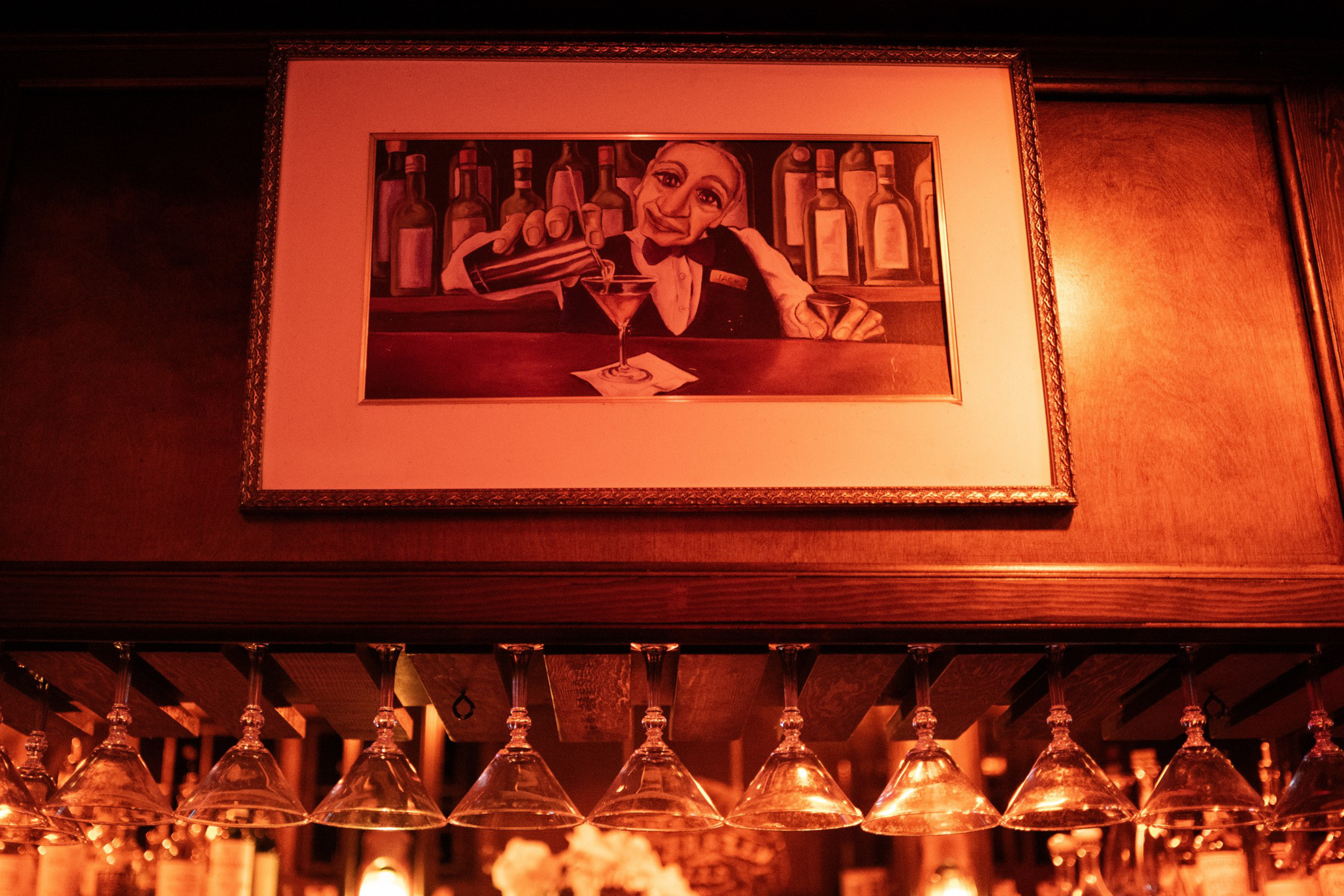 A framed painting of a bartender pouring a cocktail hangs above a rack of upside-down martini glasses in a warmly lit bar.