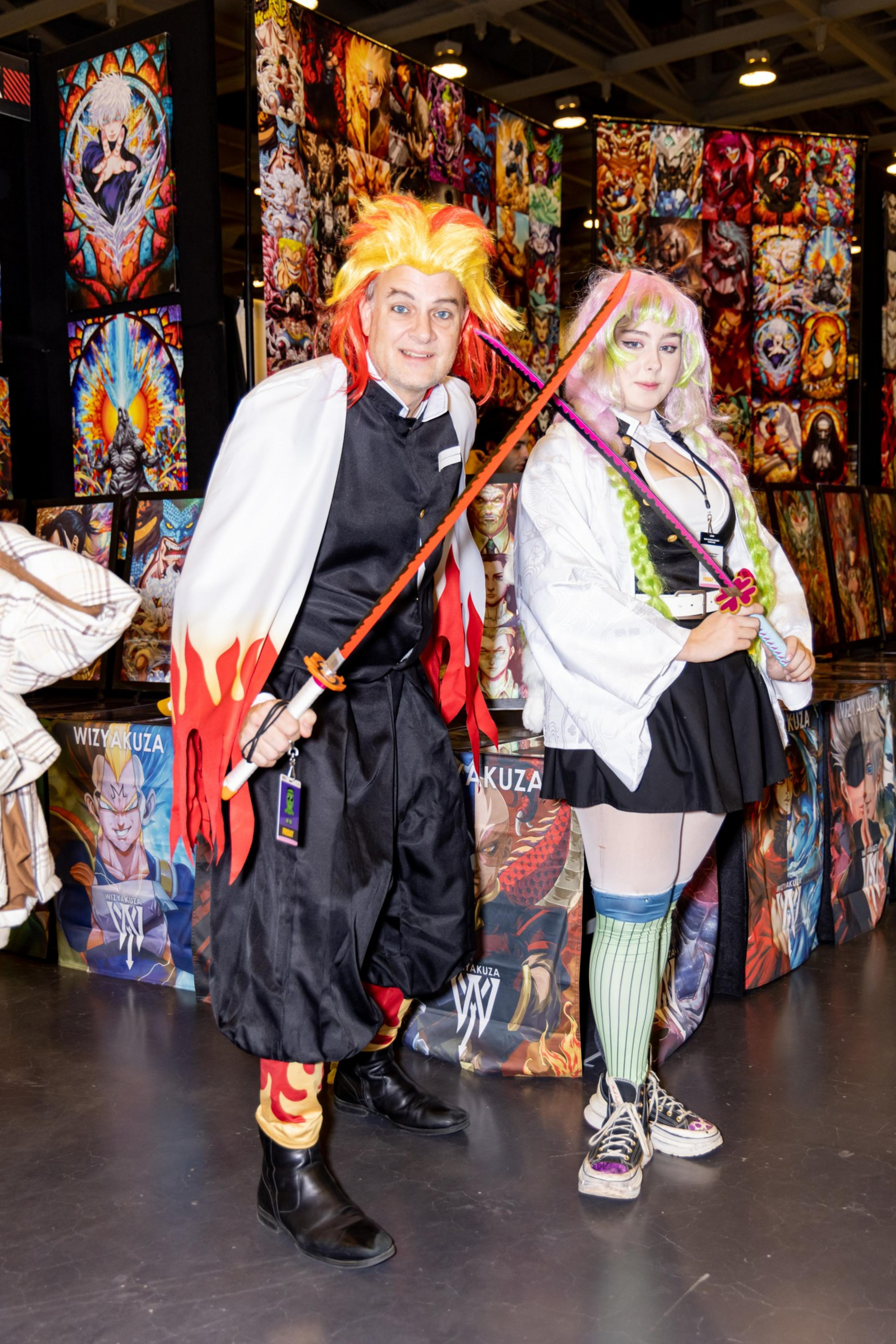 Two people are in a convention setting dressed in elaborate, colorful anime-style costumes with swords. The background is filled with vibrant posters.