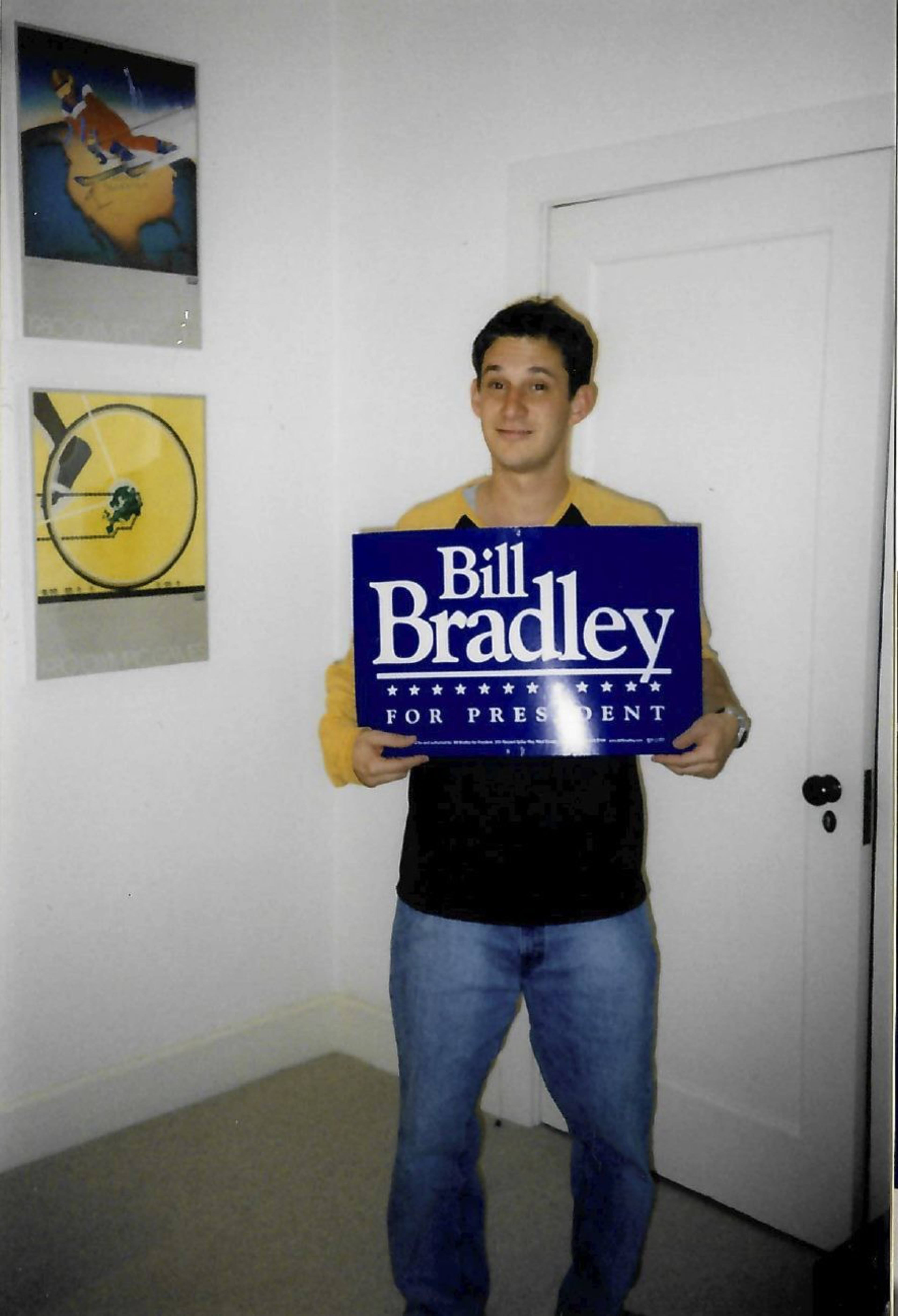A person is holding a &quot;Bill Bradley for President&quot; sign in a room with white walls and two wall posters. One poster is of a tennis racket and a ball.