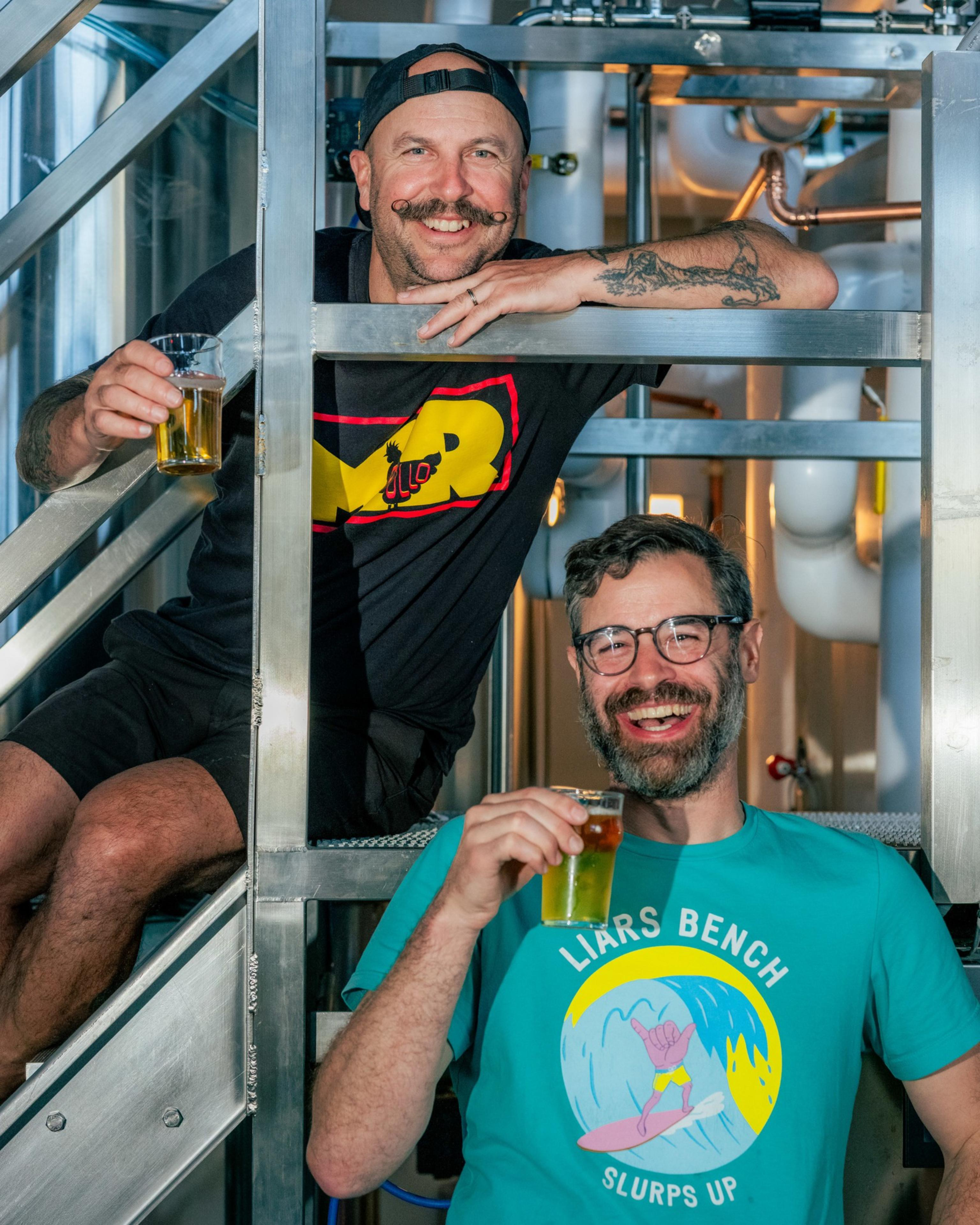 Two men are happily posing with glasses of beer in a brewery. One sits casually on a metal stair, while the other stands, both smiling warmly.