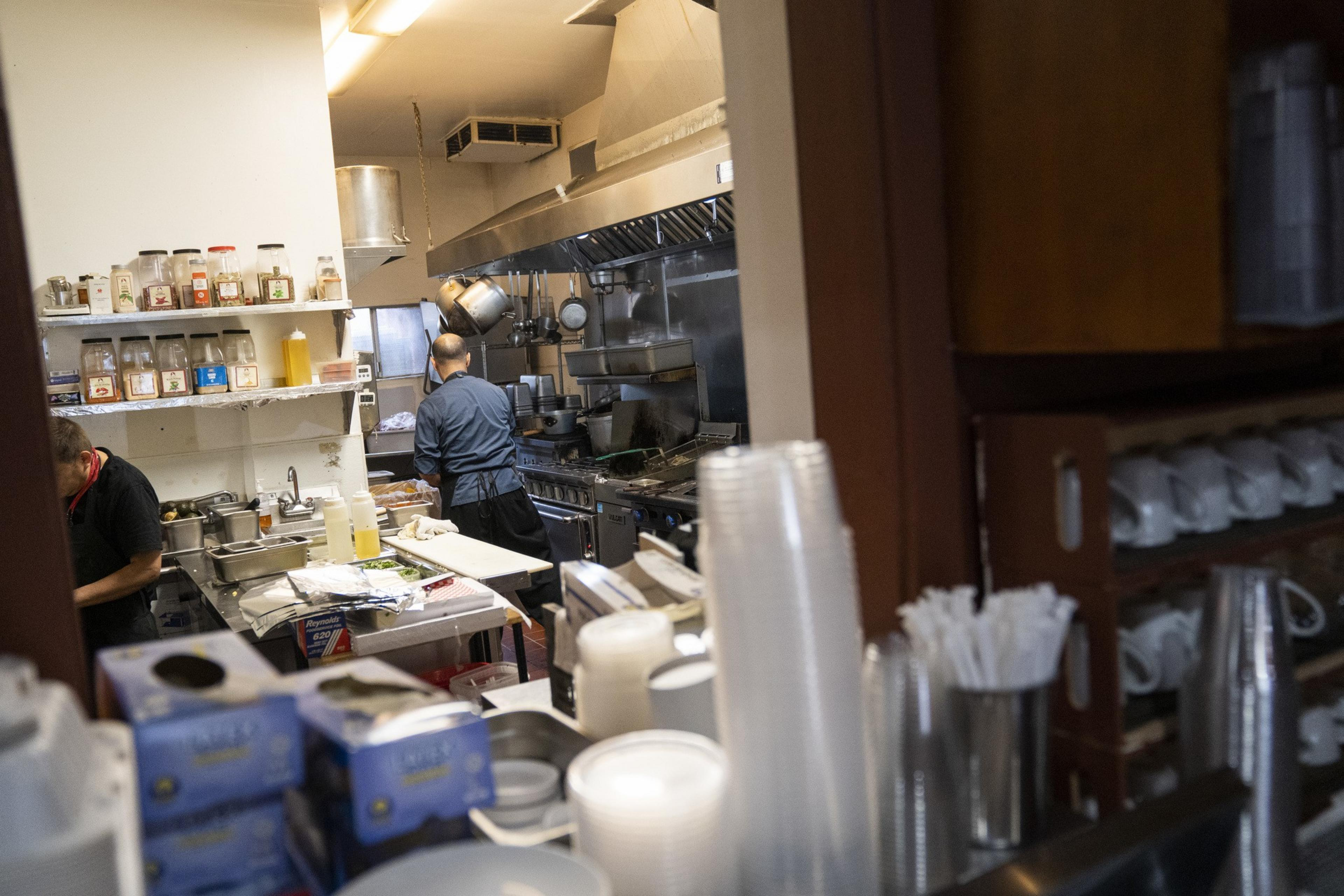 A busy restaurant kitchen with two people preparing food, shelves of spices, cooking utensils, and stacked disposable cups in the foreground.