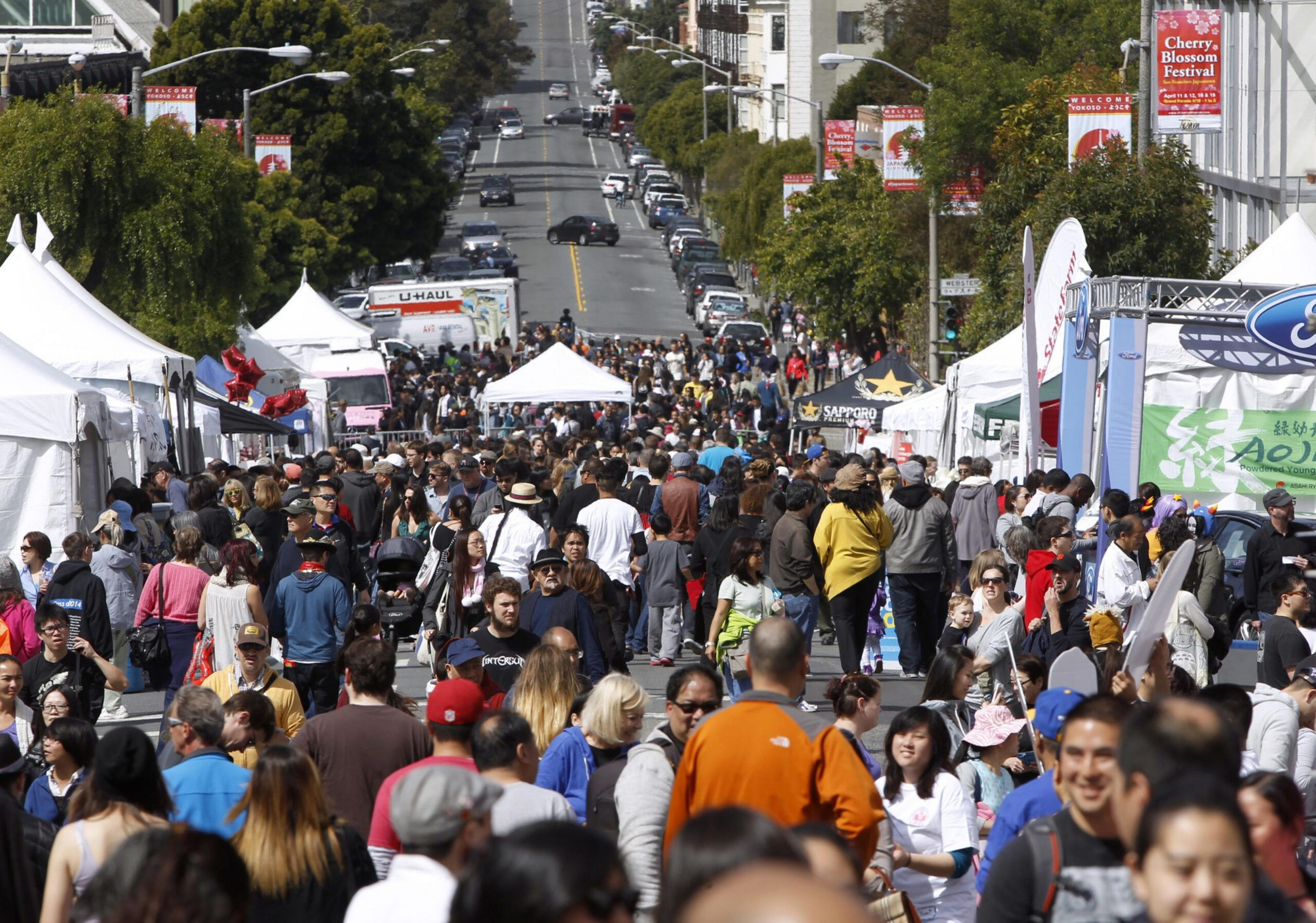 A crowded street festival with people walking among white tents, booths, and banners advertising a Cherry Blossom Festival.