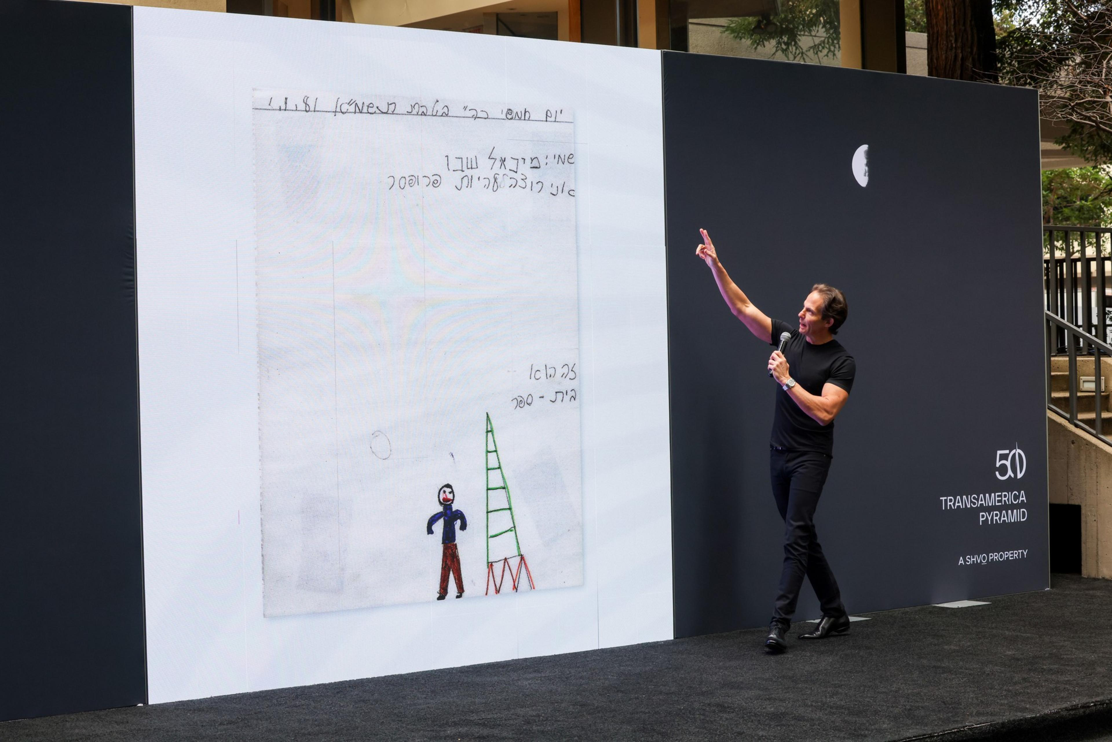 A man in black clothes holds a microphone and points at a large screen displaying a child-like drawing of a person and a green ladder.