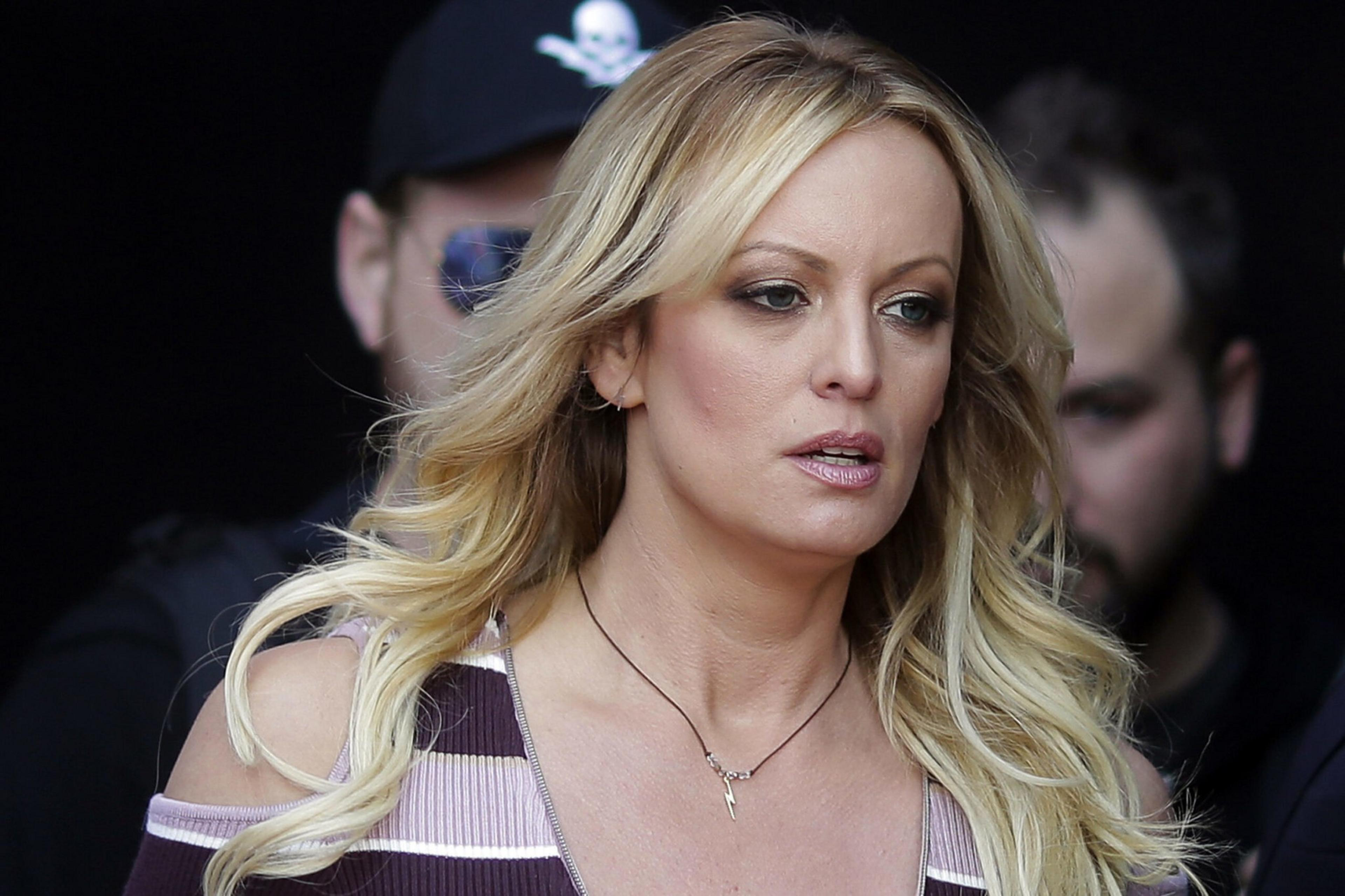 A blonde woman with wavy hair wears a striped top and a necklace, walking past two men in the background, one with a pirate hat and sunglasses.
