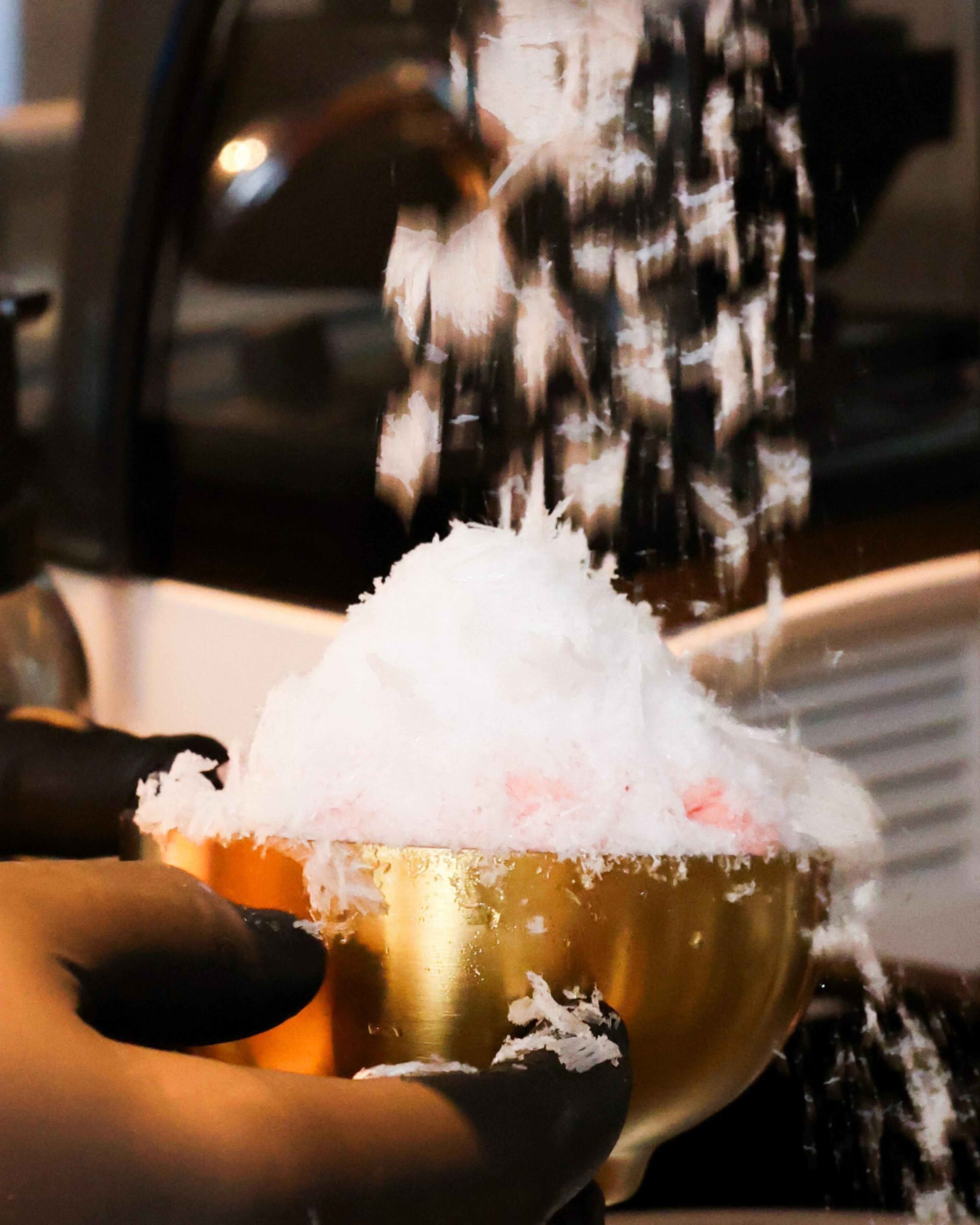 A pair of gloved hands holds a golden bowl filled with fluffy, white shaved ice, with more ice being sprinkled from above.