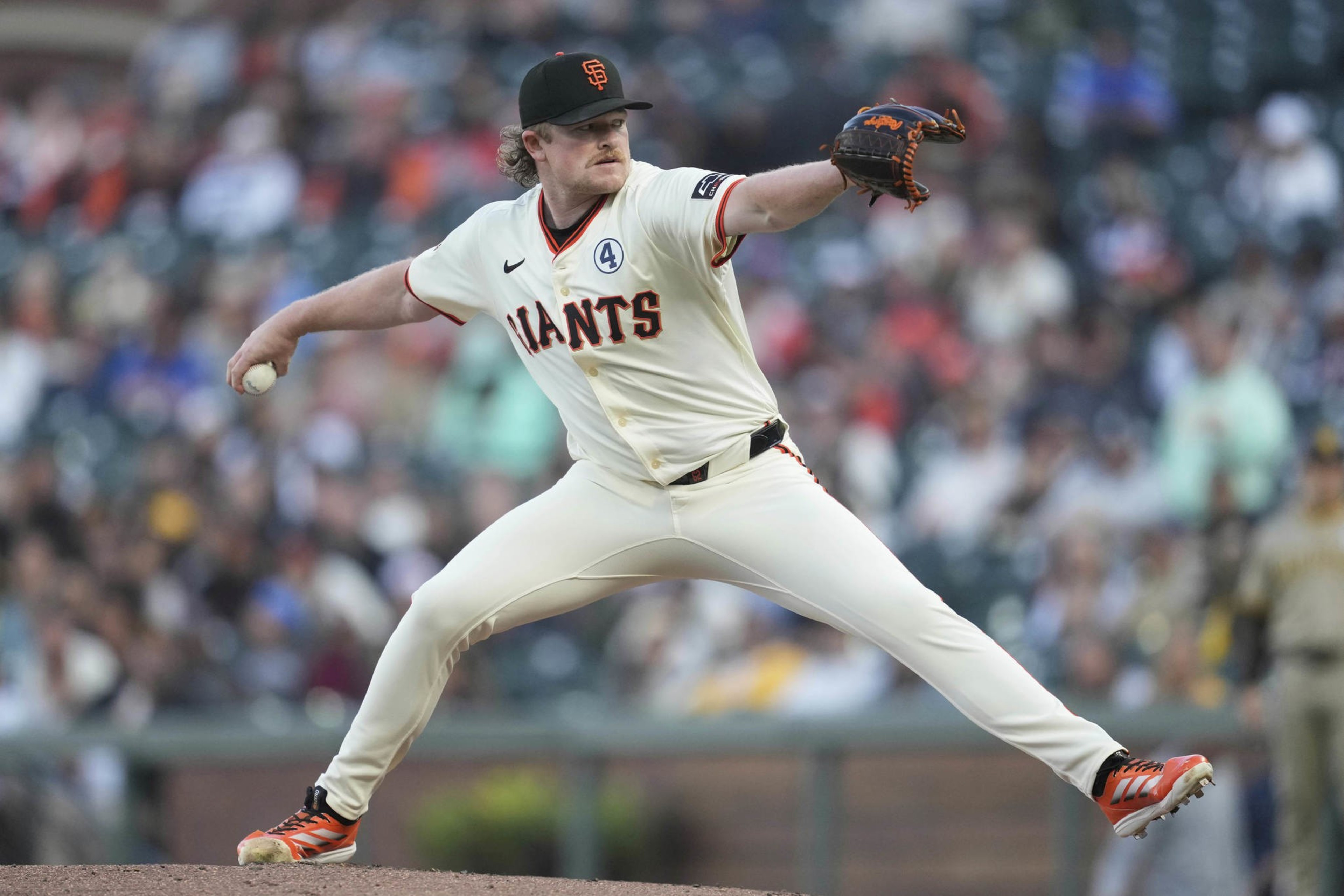 A baseball player in a white &quot;Giants&quot; uniform is pitching on the mound, wearing an SF cap and orange cleats, with a blurred crowd in the background.