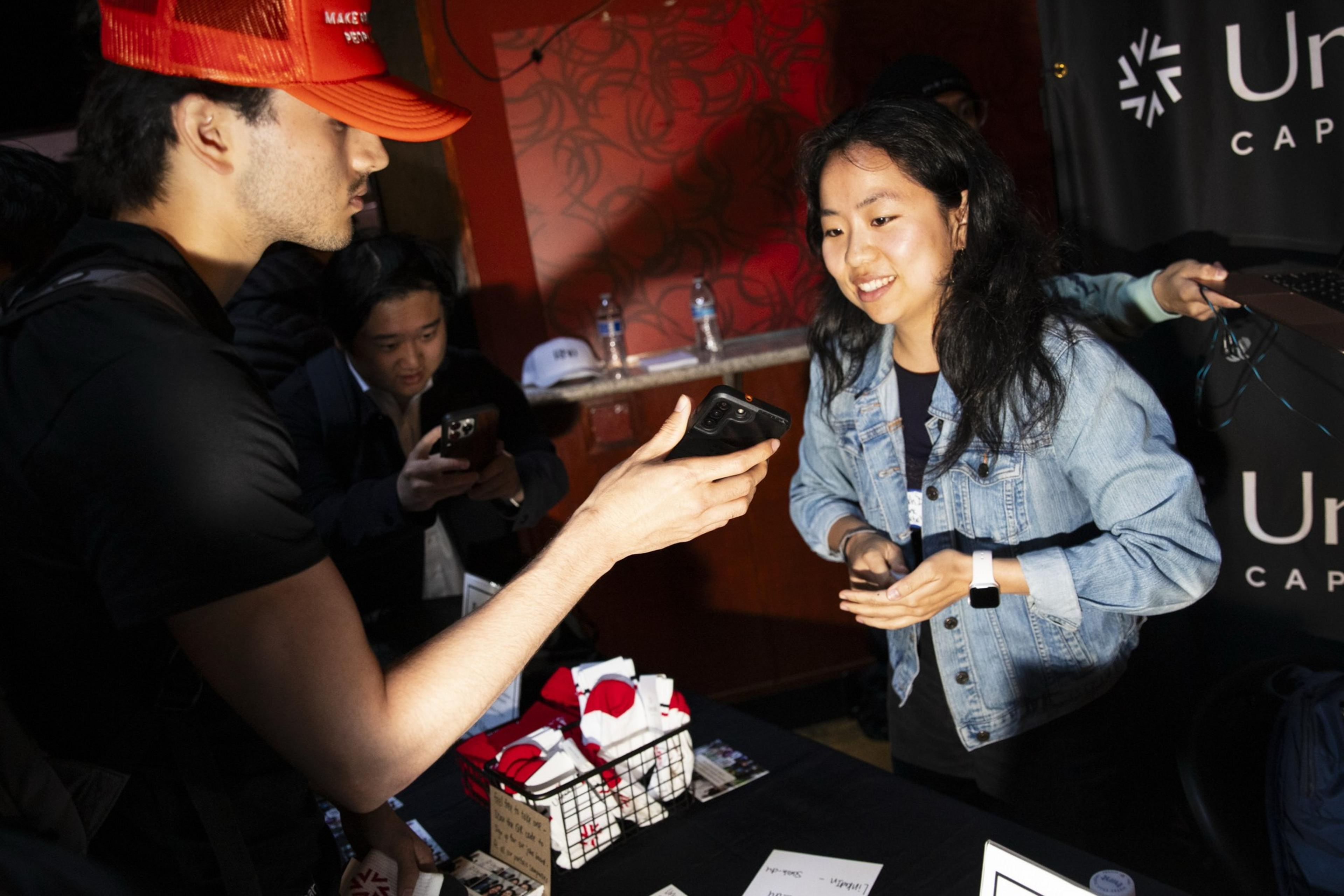 A man in a red hat hands a phone to a smiling woman in a denim jacket across a table with items on it. Another person uses a phone in the background.
