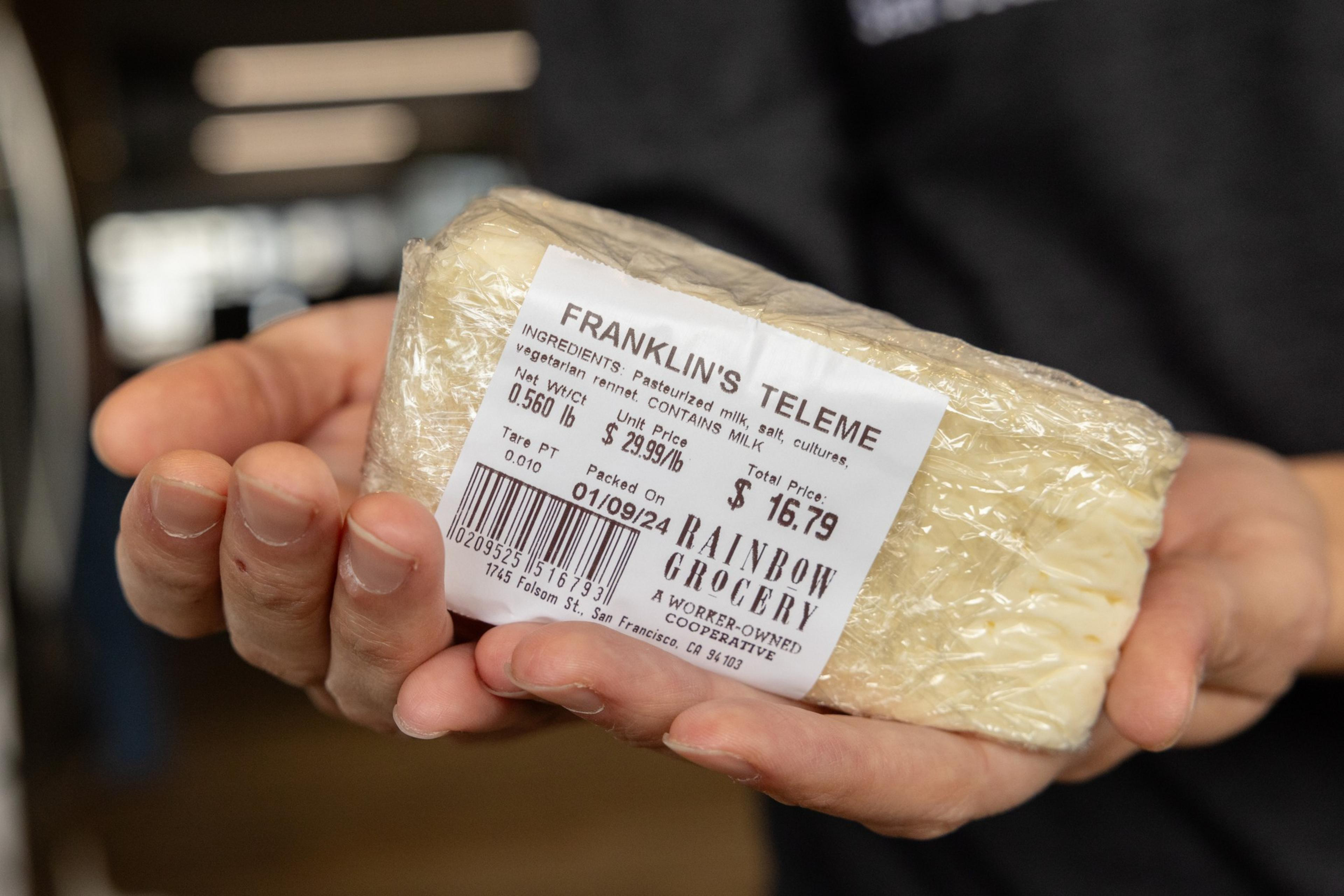 A person is holding a wrapped block of Franklin's Teleme cheese with a price label.