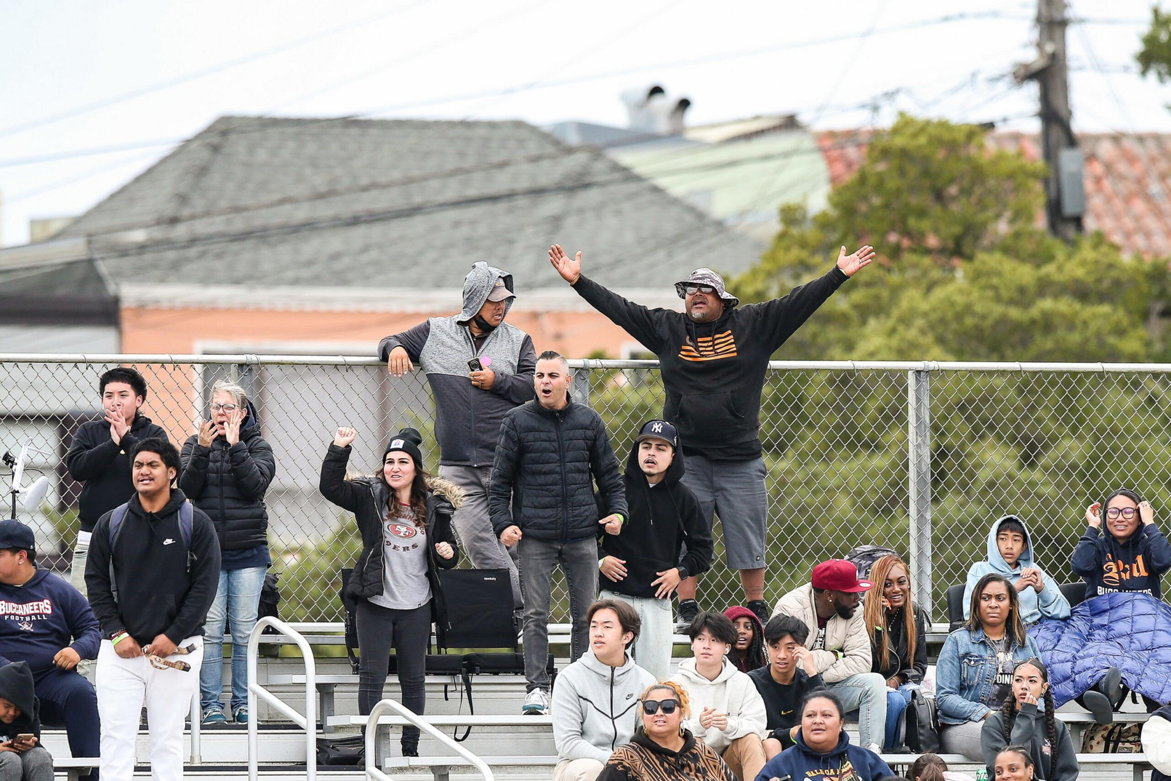 A group of diverse spectators in casual clothing cheer and watch an event, some standing with raised arms, others sitting on metal bleachers outdoors.