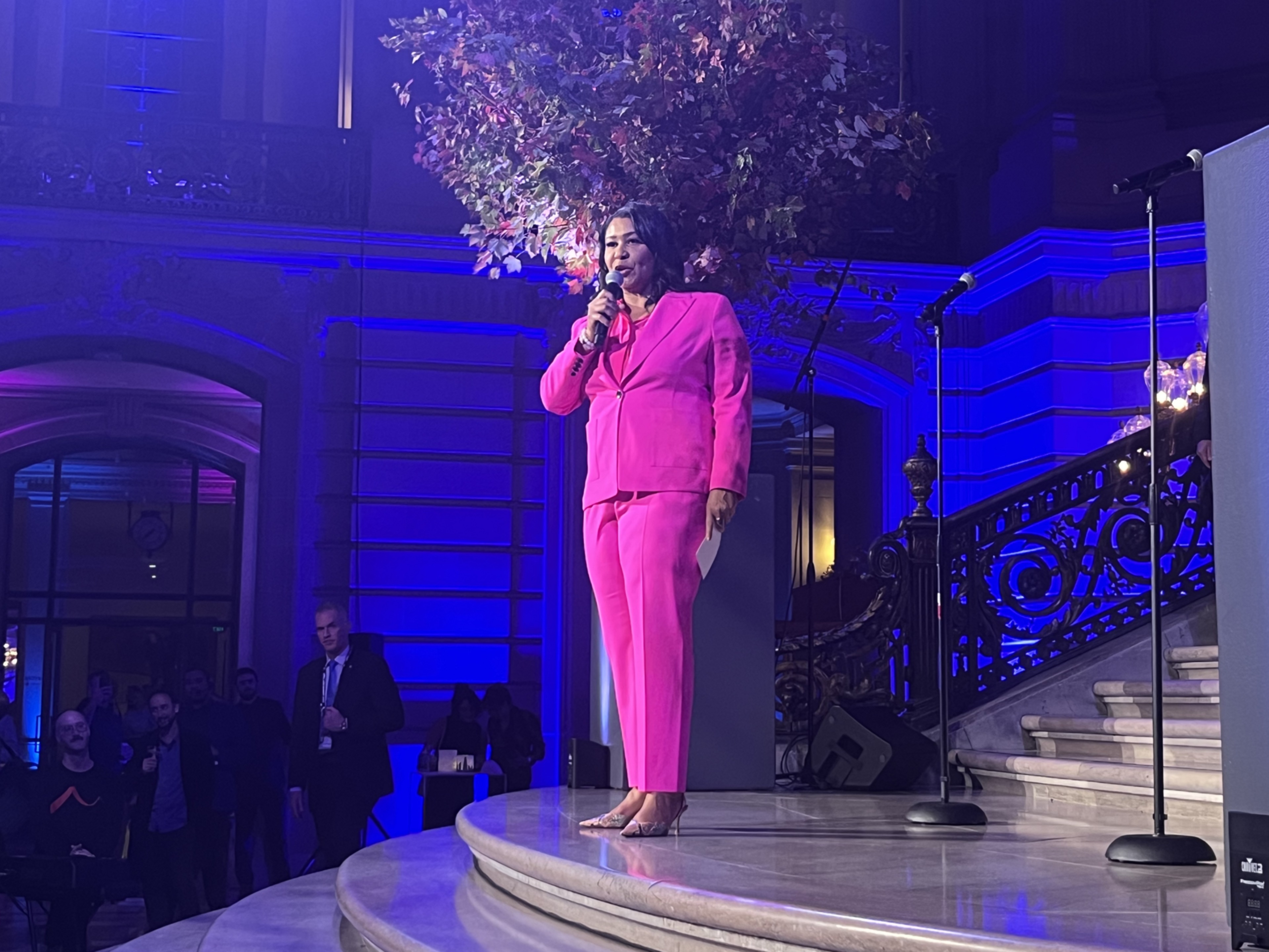 A woman in a pink suit addresses a crowd with a microphone.