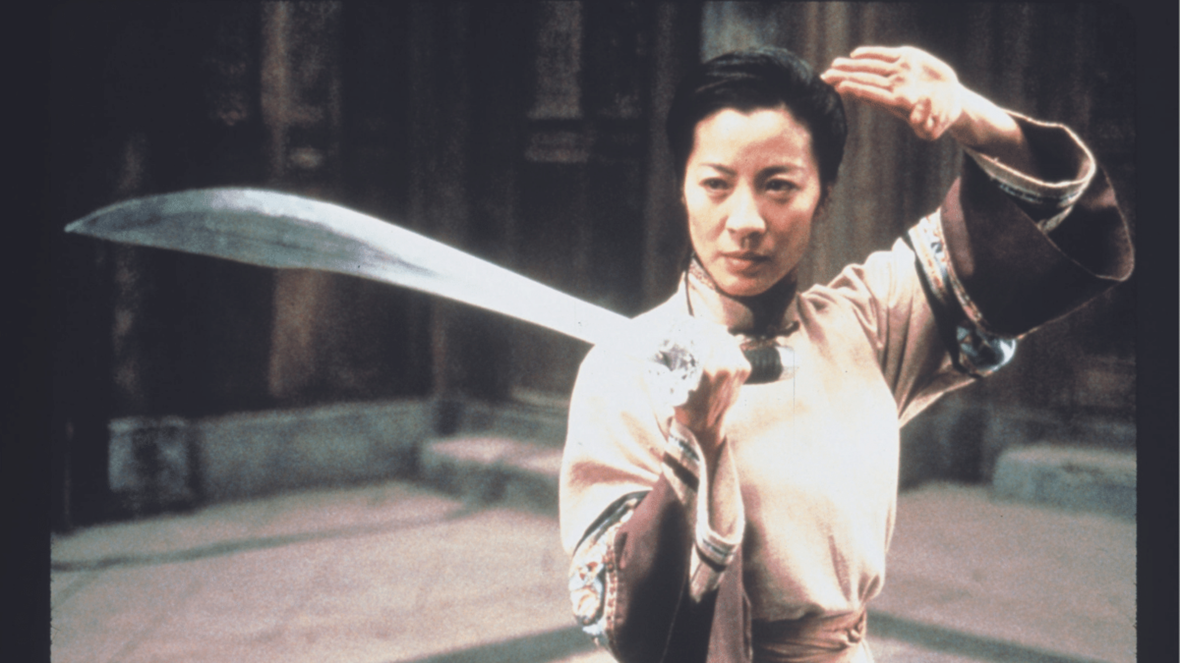 A woman in traditional clothing holds a sword upright with one hand while raising her other hand near her head in a martial arts stance.
