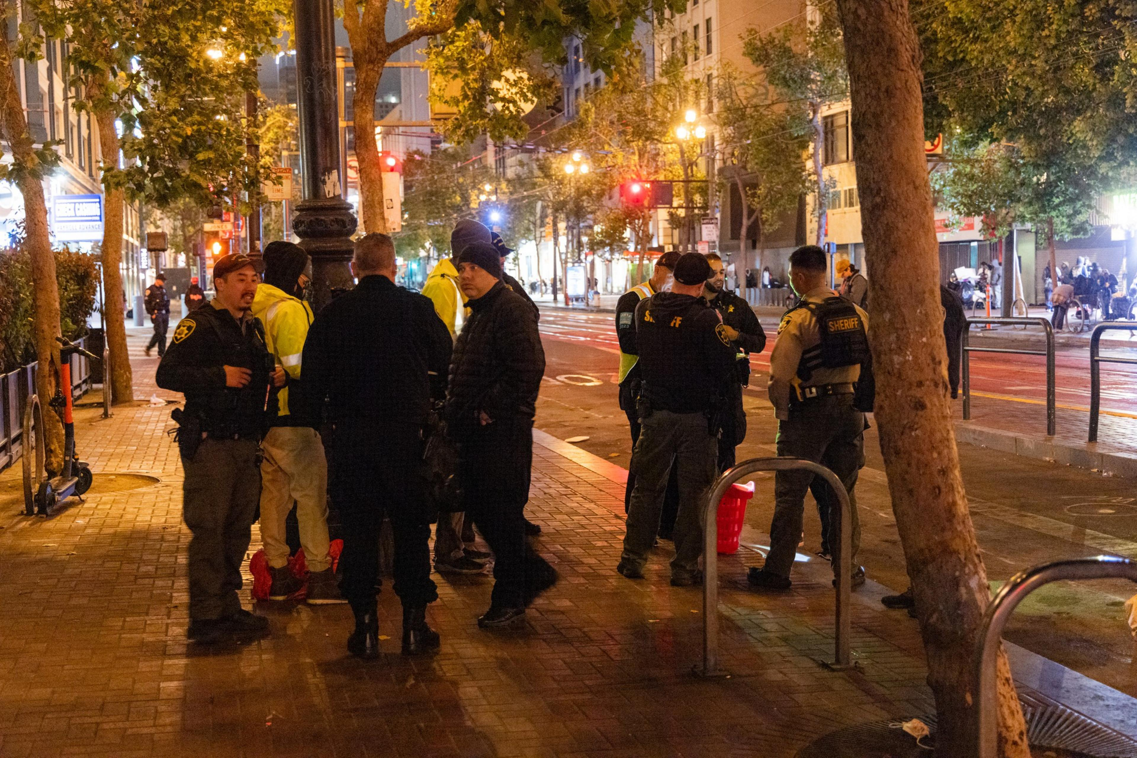 A group of law enforcement officers and individuals stand gathered on a city sidewalk at night, with trees and streetlights illuminating the scene.