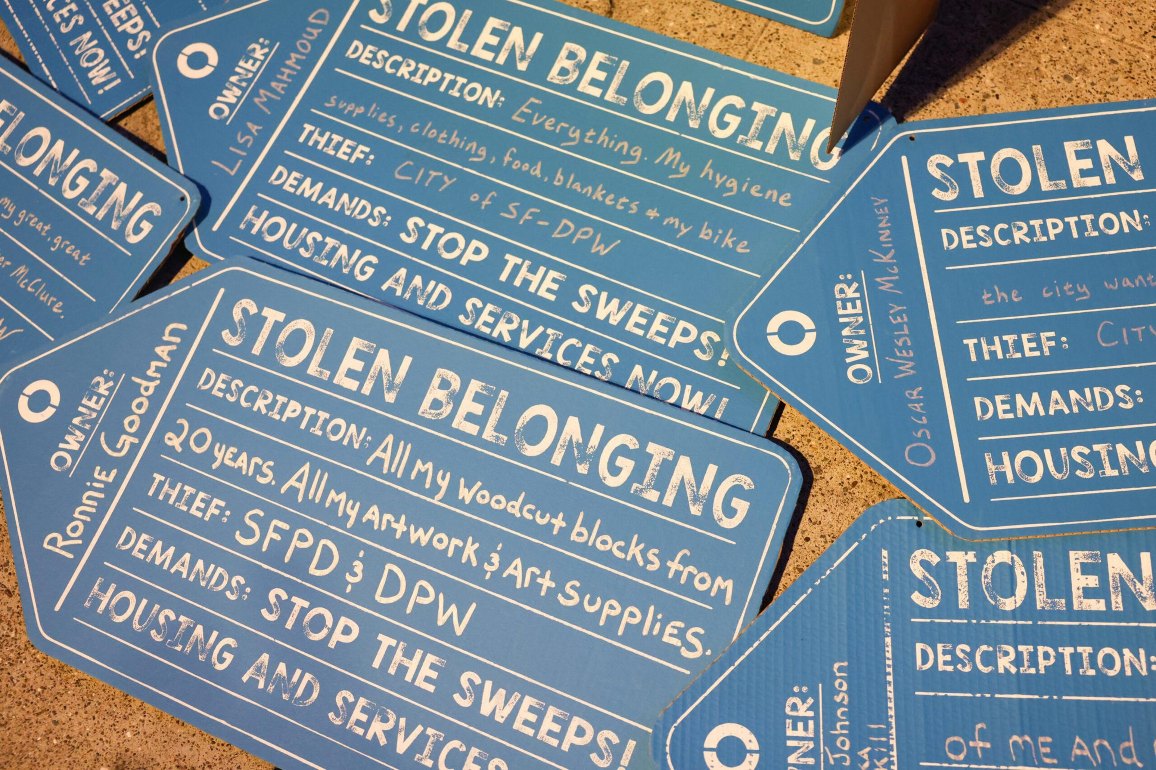 Blue signs labeled “STOLEN BELONGING” detail stolen personal items, the thieves (city agencies), and demands such as stopping sweeps and providing housing.