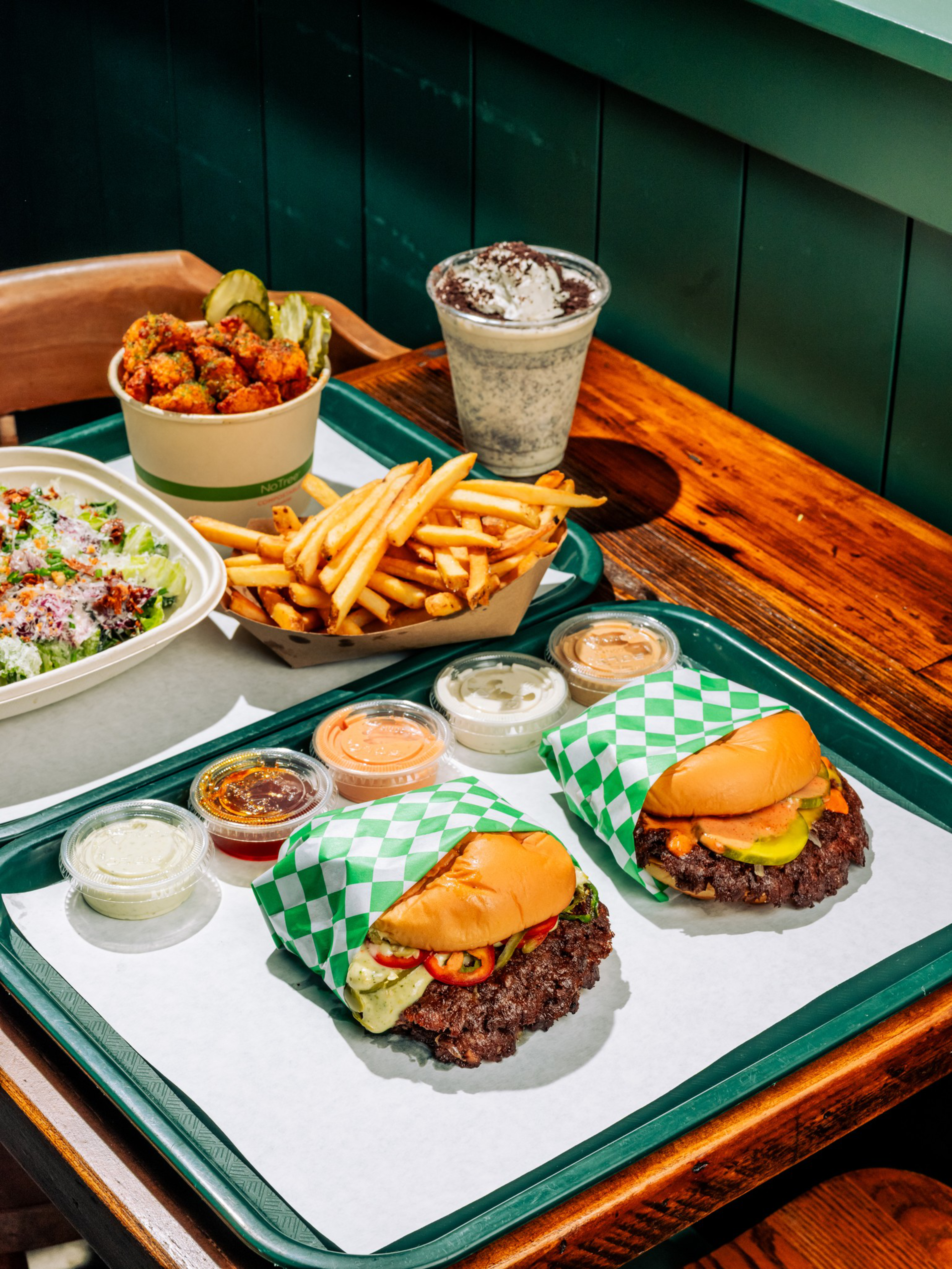 Two burgers wrapped in green and white checkered paper, a tray of French fries, a cup of fried snacks with pickles, a salad, multiple dipping sauces, and a chocolate milkshake.