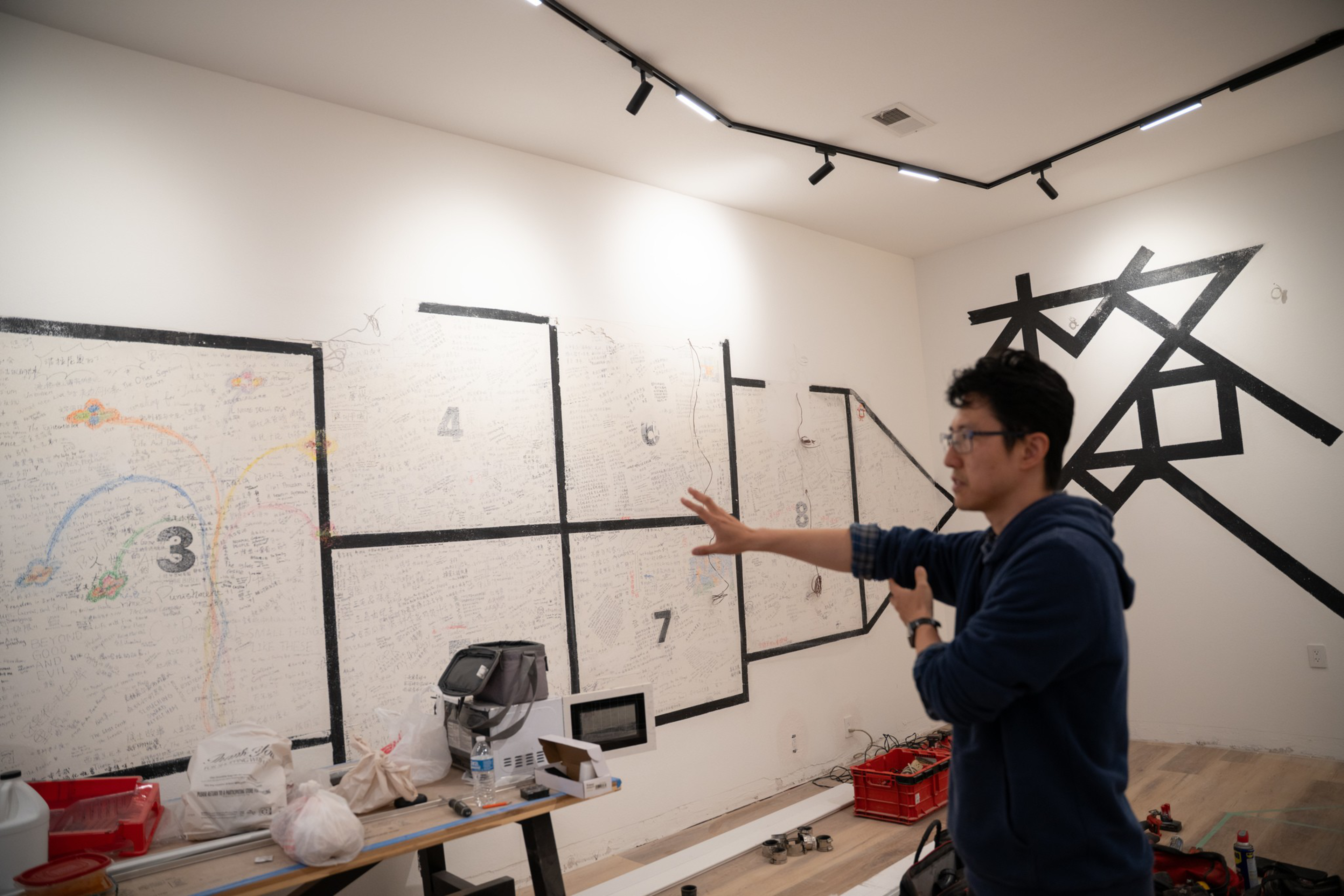 A person wearing glasses and a blue hoodie gestures toward a white wall marked with numbered sections and handwritten notes, with some items on a nearby table.