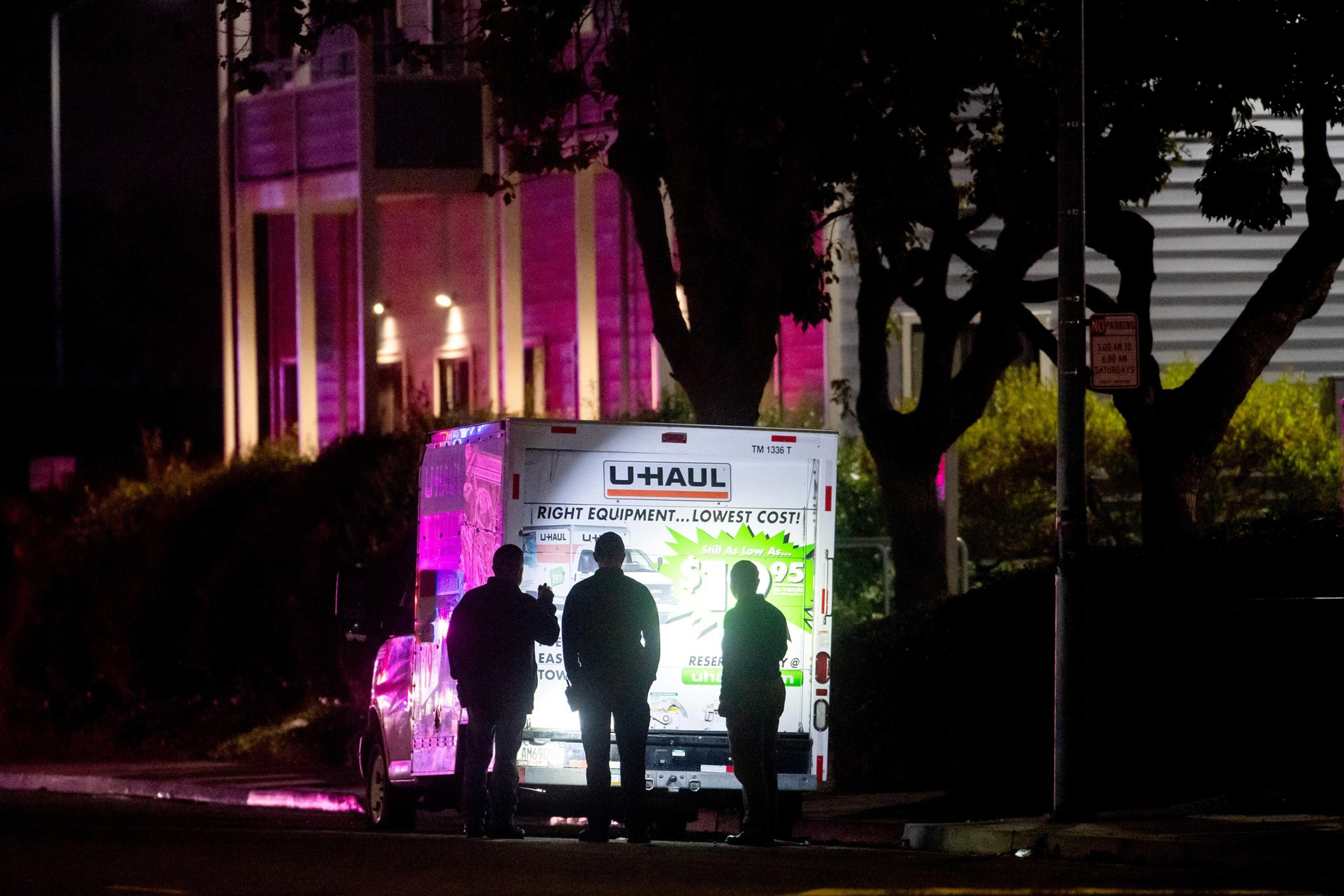 Three people stand behind a U-Haul truck at night, with bright lights illuminating the truck and casting purple hues on nearby buildings.