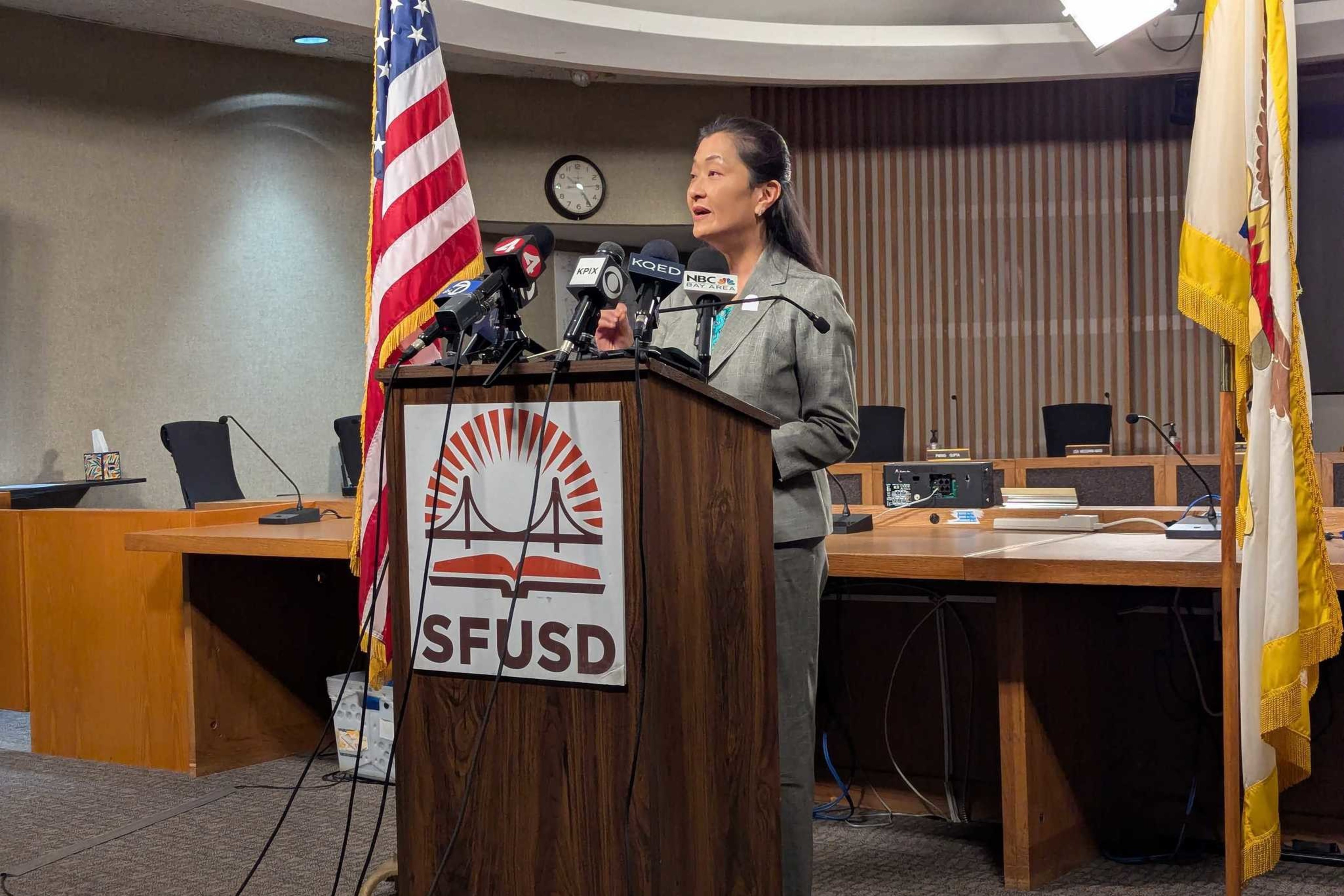 A woman in a gray suit speaks at an SFUSD podium with multiple microphones, flanked by the American flag and another flag inside a boardroom.