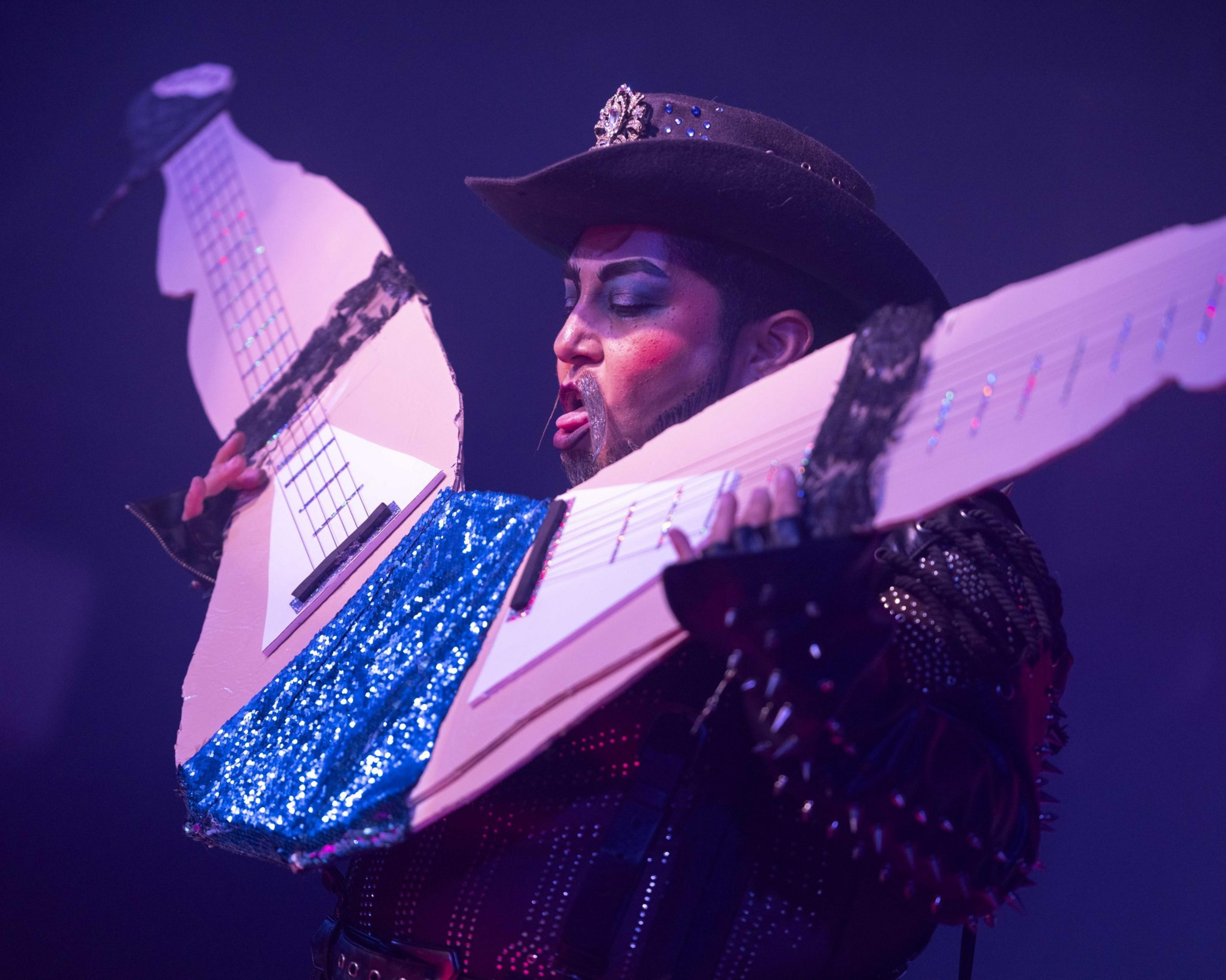 A performer in dramatic makeup and a studded outfit holds an oversized, double-neck cardboard guitar decorated with blue sequins, appearing mid-act on stage.