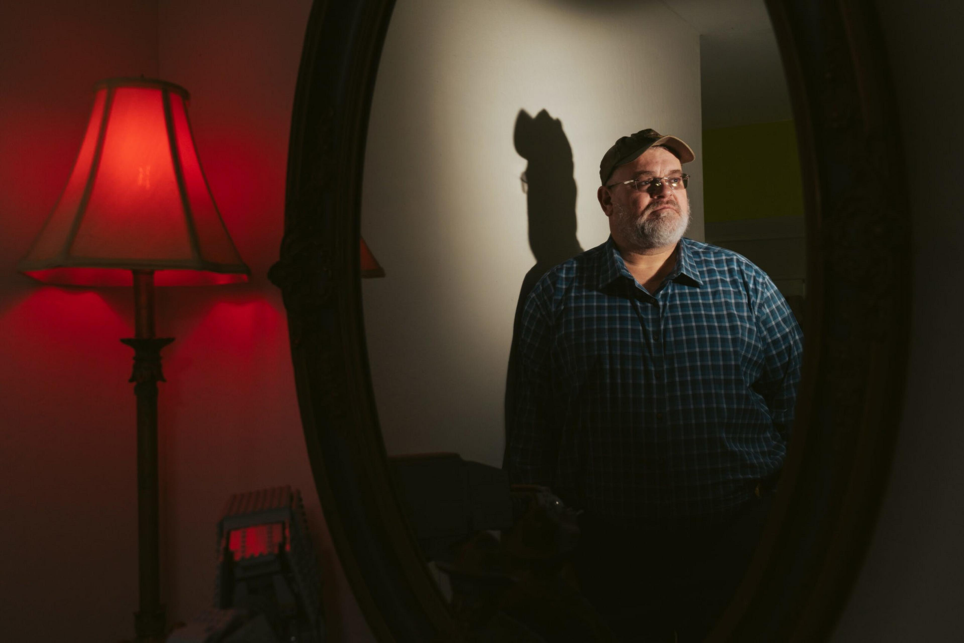 A man wearing glasses, a cap, and a blue plaid shirt stands looking to the side, reflected in a mirror with a red-lit lamp nearby.