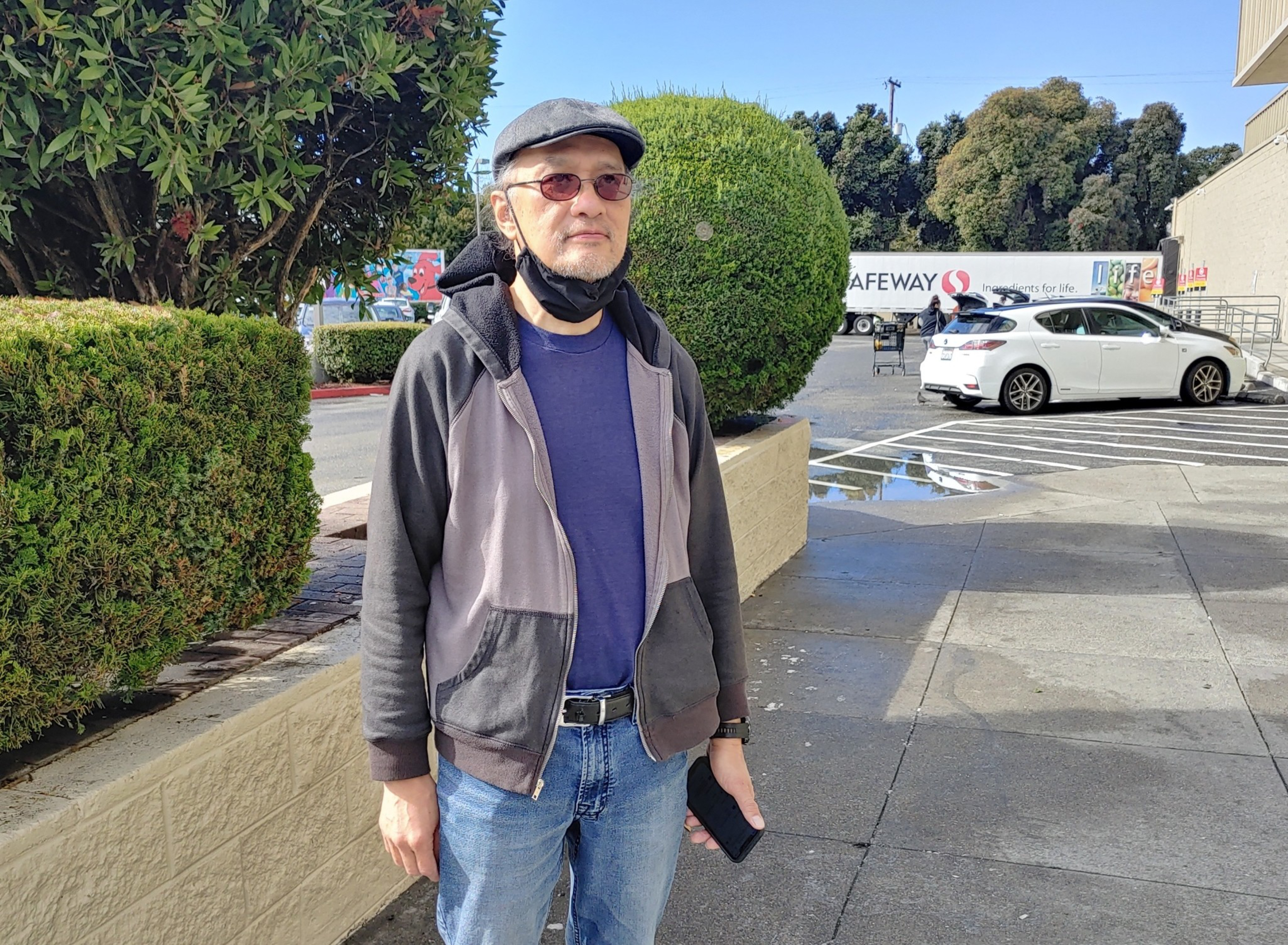 A man wearing a cap, sunglasses, and a face mask under his chin stands near trimmed bushes in a parking lot with cars and a Safeway truck in the background.