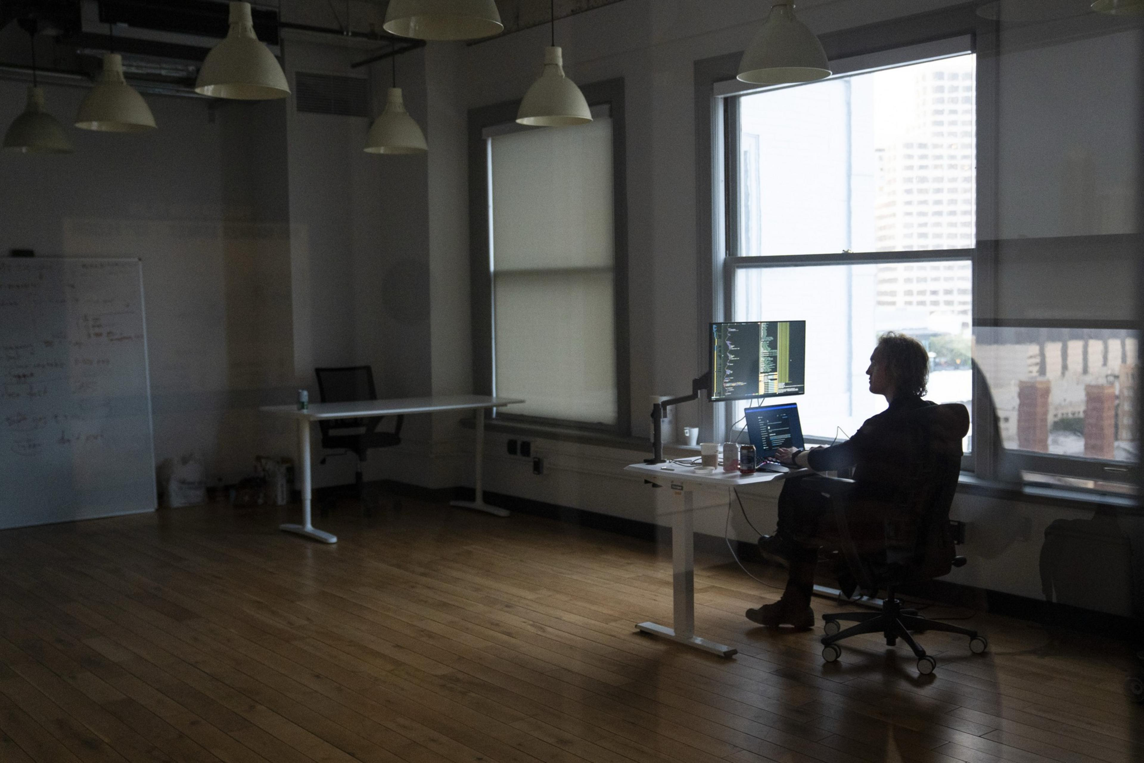 A person works at a desk in a dimly lit office, typing on a laptop with a monitor displaying code. Large windows and various lights illuminate the space.