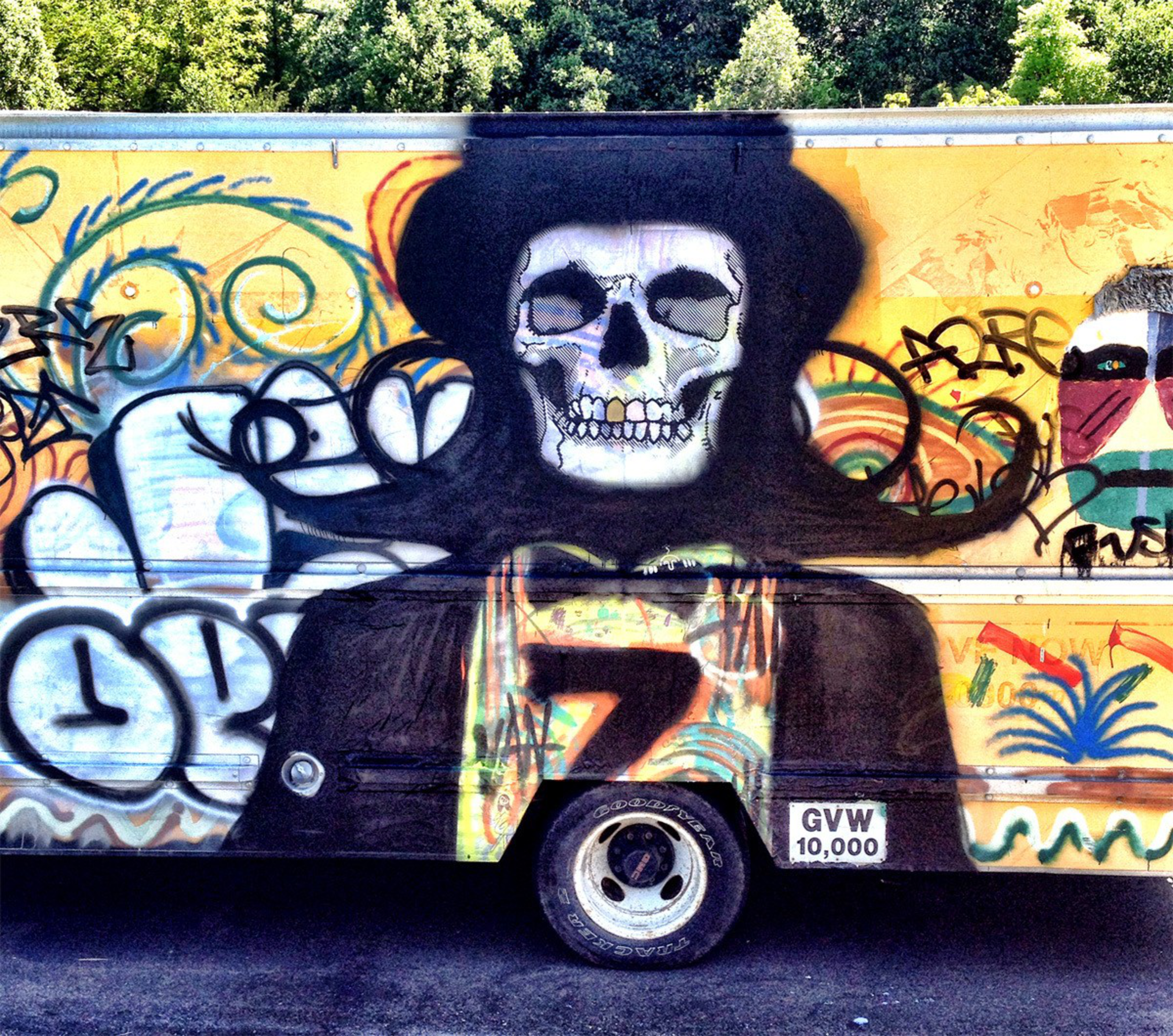 A graffiti mural of a skull wearing a wide-brimmed black hat and cape adorns a yellow vehicle side with colorful swirls and abstract patterns.