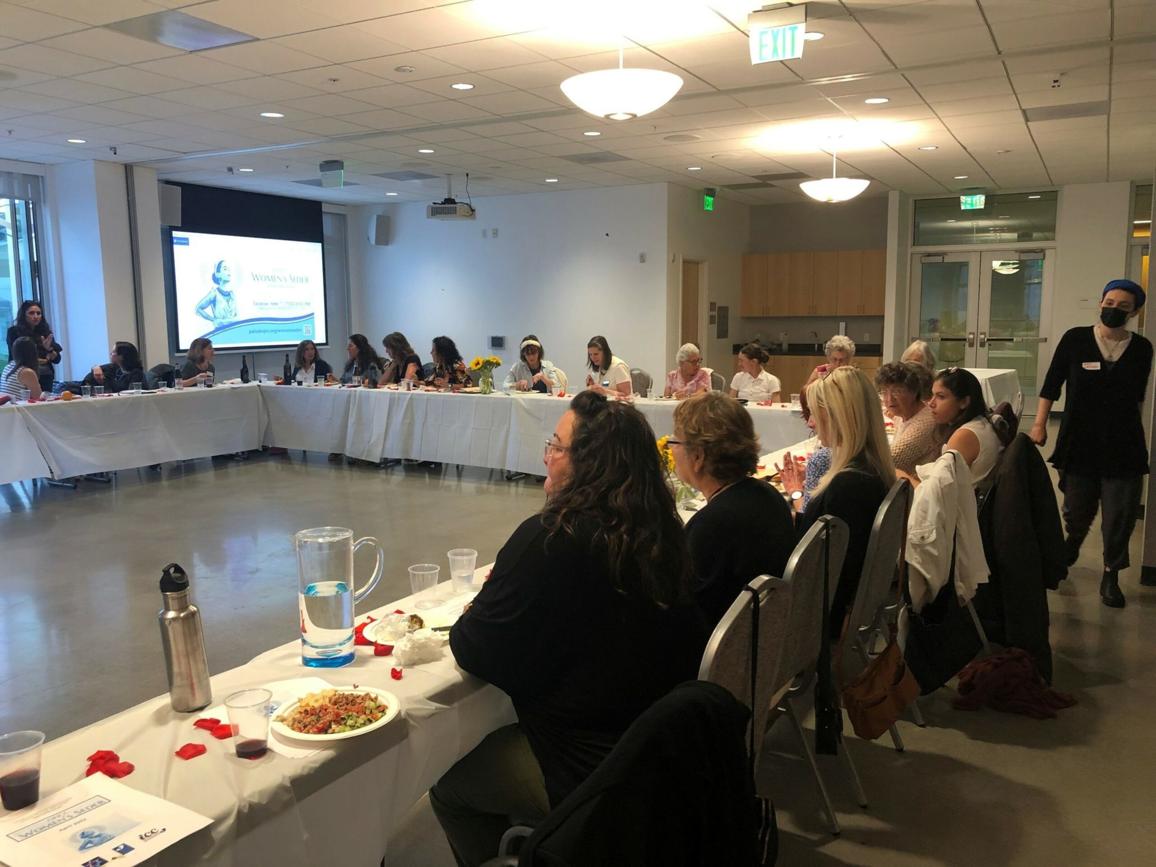 A group of people sit around U-shaped tables covered with white cloths, listening to a speaker near a projection screen in a brightly lit conference room.