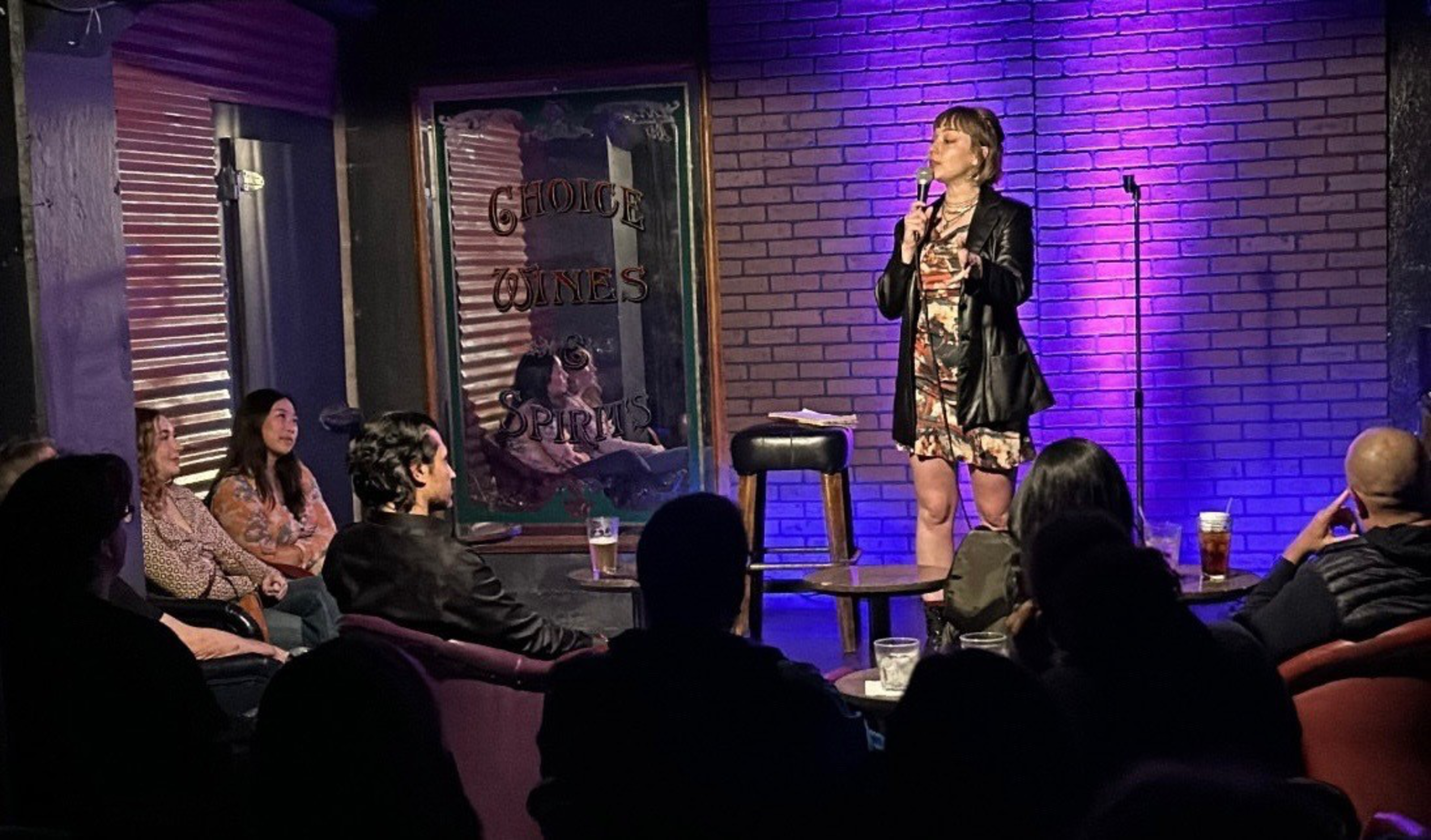 A woman in a floral dress and black jacket performs stand-up comedy with a microphone on stage, facing an attentive seated audience in a dimly lit club.
