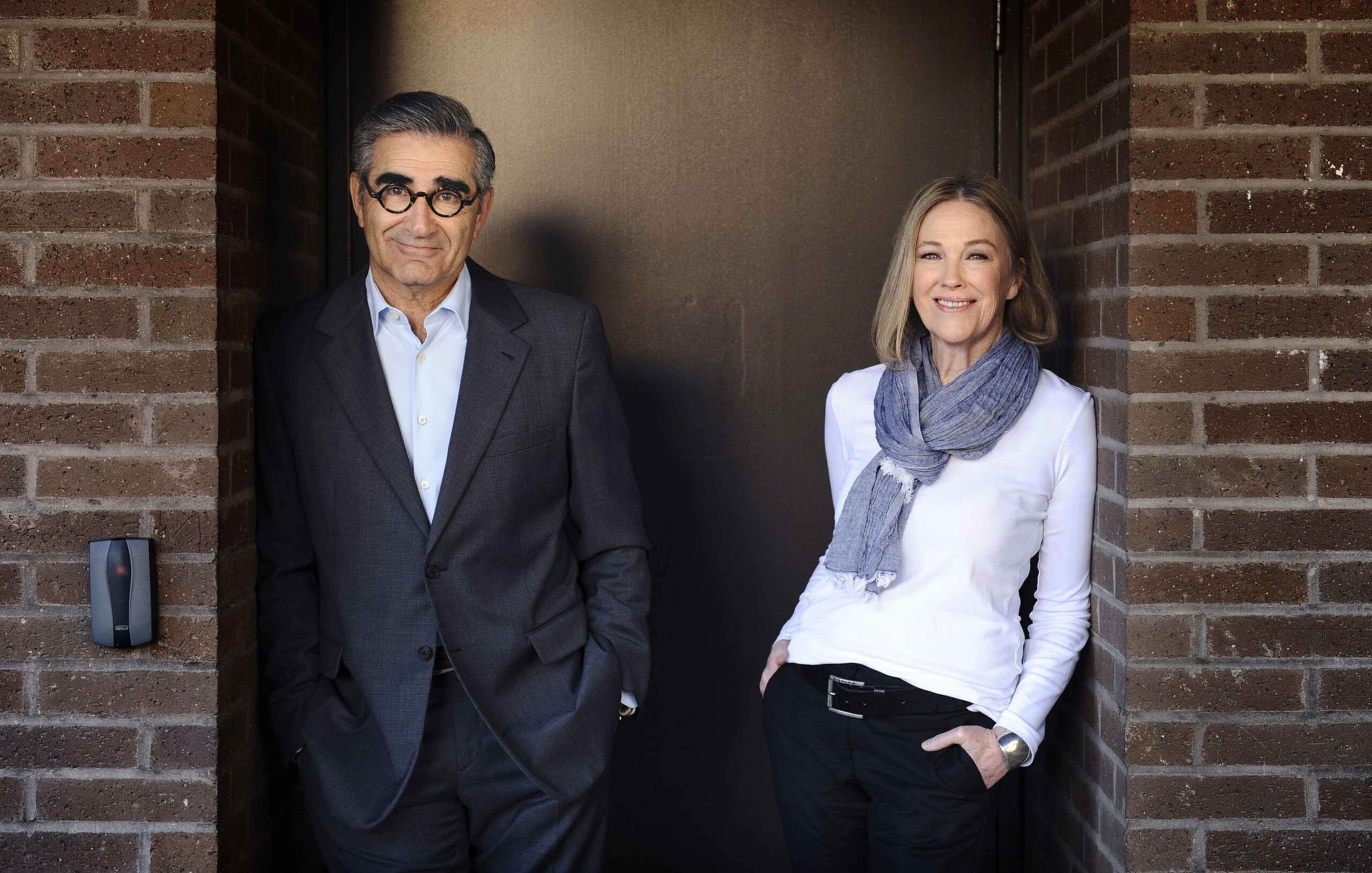 A man in a dark suit and glasses and a woman in a white top with a gray scarf stand casually against a brick wall facing the camera.