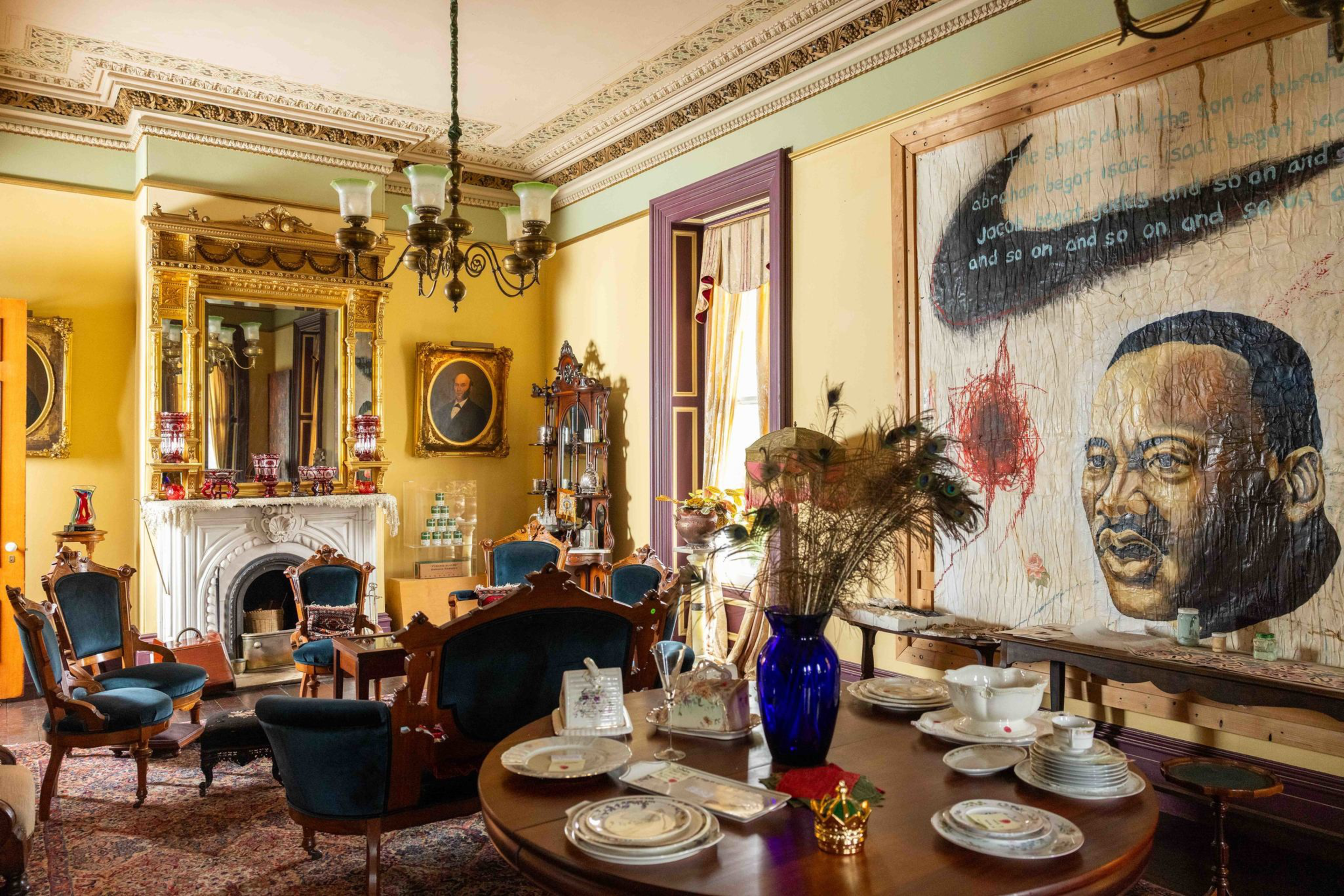 A vintage room with blue velvet chairs, a white fireplace, a large painted portrait of Martin Luther King Jr., antique furniture, and a chandelier.