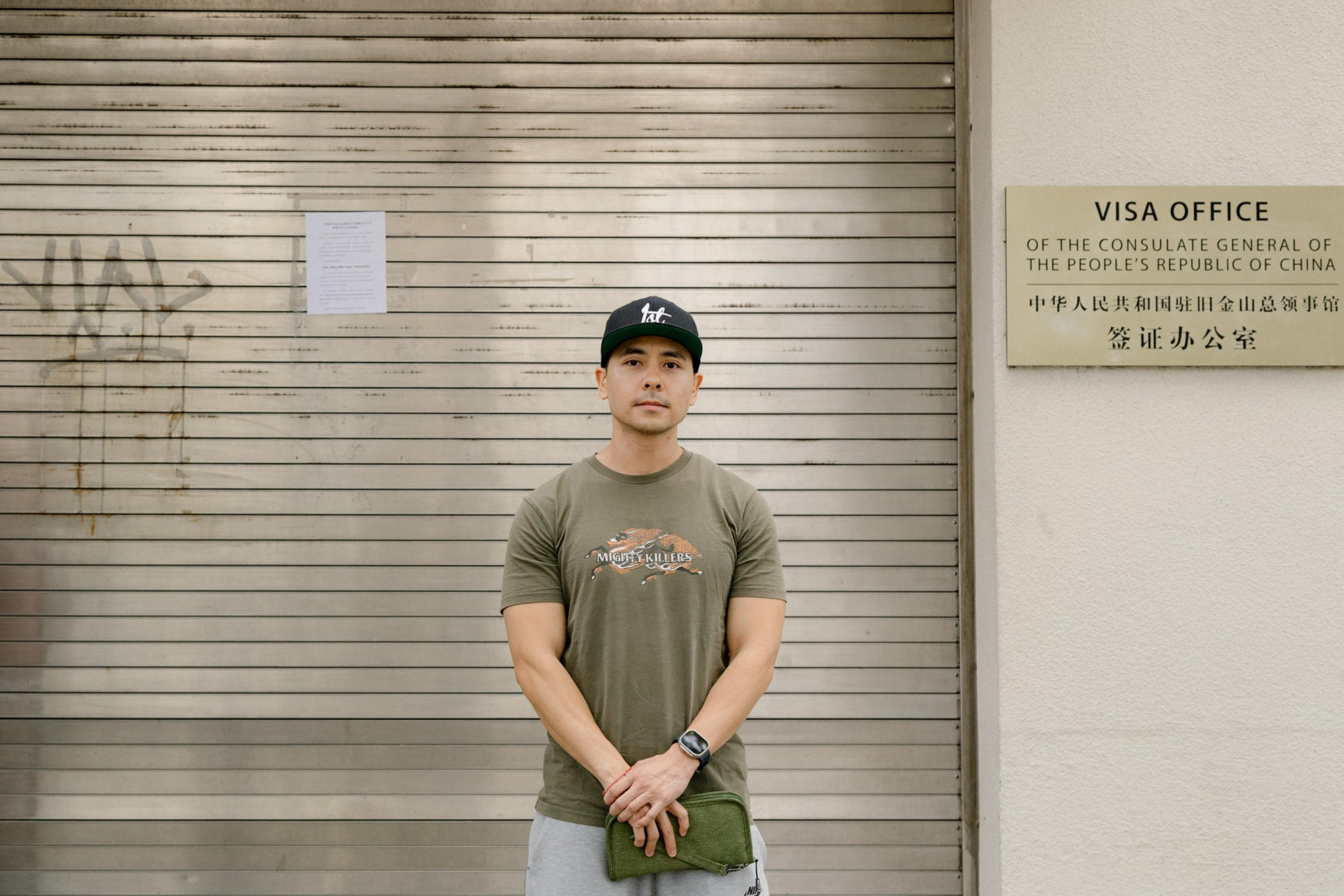 A man wearing a green "Mighty Killers" t-shirt and black cap stands with clasped hands in front of a closed metal shutter next to a Visa Office sign.