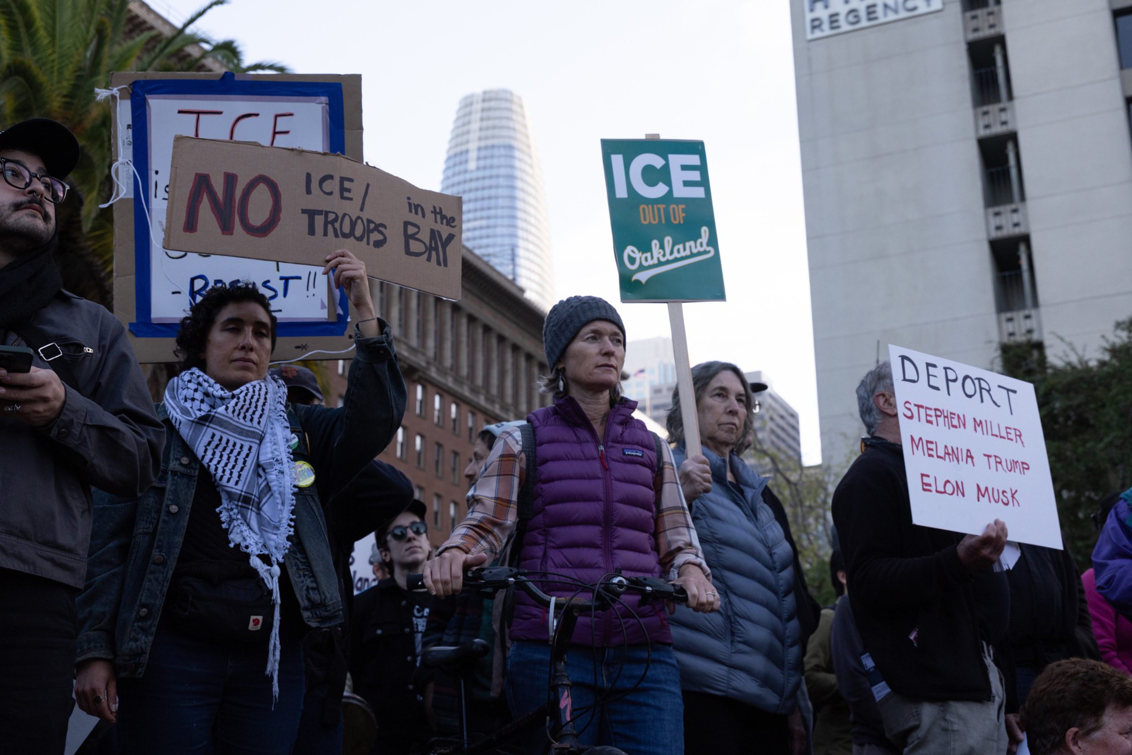 People hold signs protesting ICE presence, with messages like “NO ICE/TROOPS in the BAY,” “ICE OUT OF Oakland,” and “DEPORT Stephen Miller, Melania Trump, Elon Musk.”