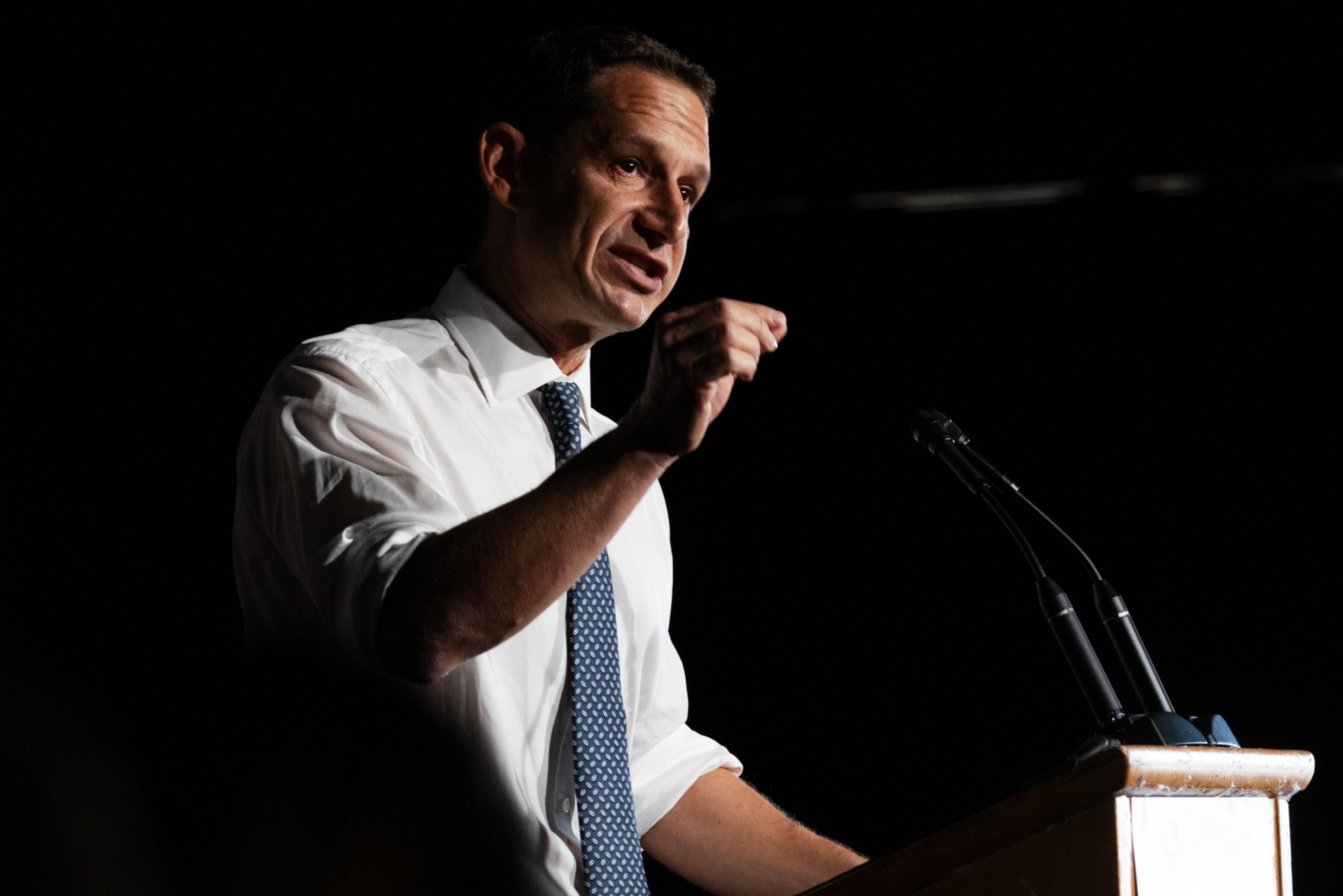 Daniel Lurie in a white shirt and blue tie speaks passionately at a podium with two microphones, gesturing with his right hand against a dark background.