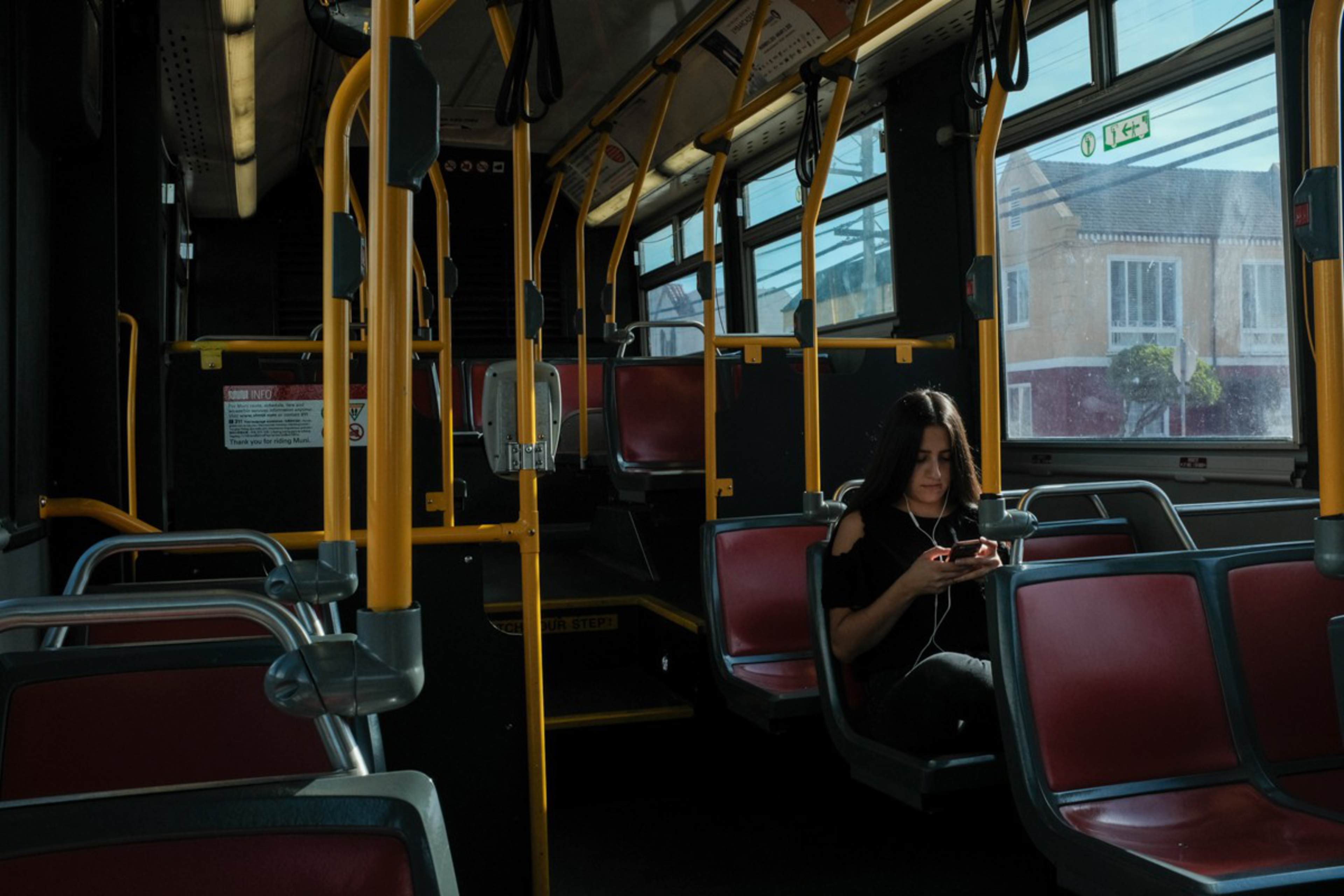 A woman with long dark hair sits alone on a bus, wearing earphones and looking at her phone, with sunlight streaming through the windows.