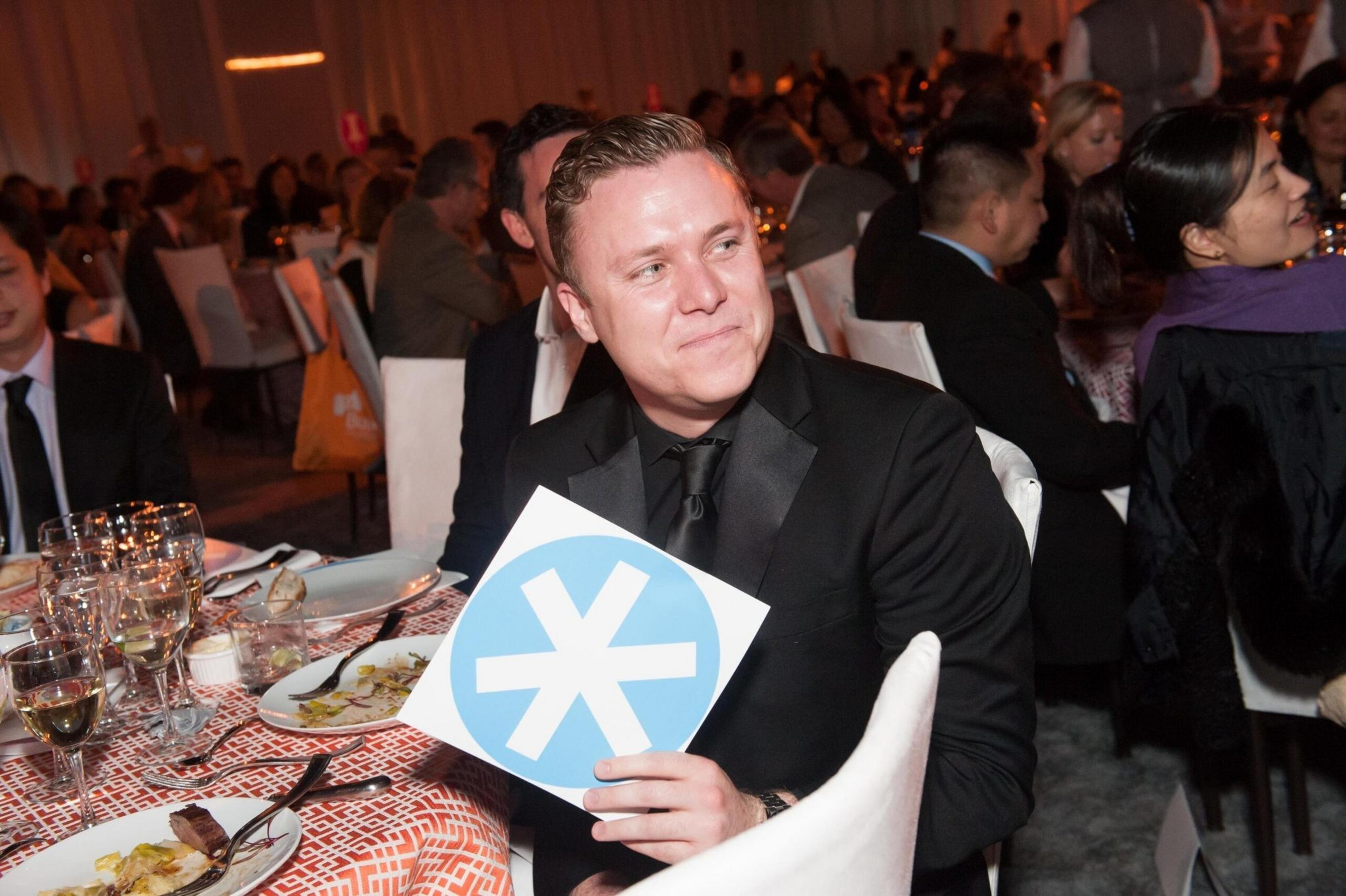 A man in a suit holds a blue sign with a white asterisk at a crowded, formal dining event. There's food and wine on the table in front of him.