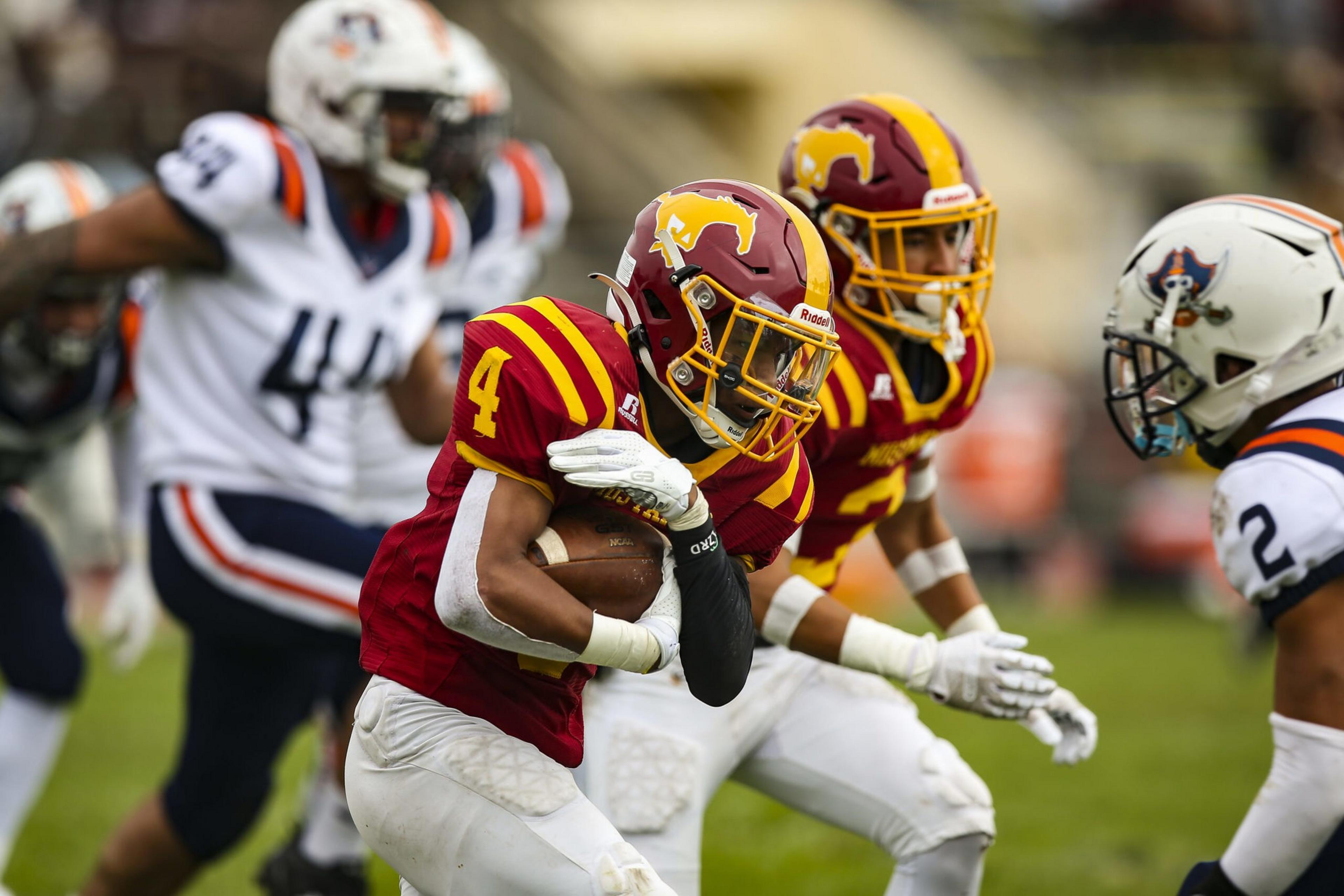 A football player in a red and yellow uniform clutches the ball, guarded by a teammate, as two opposing players in white uniforms approach.